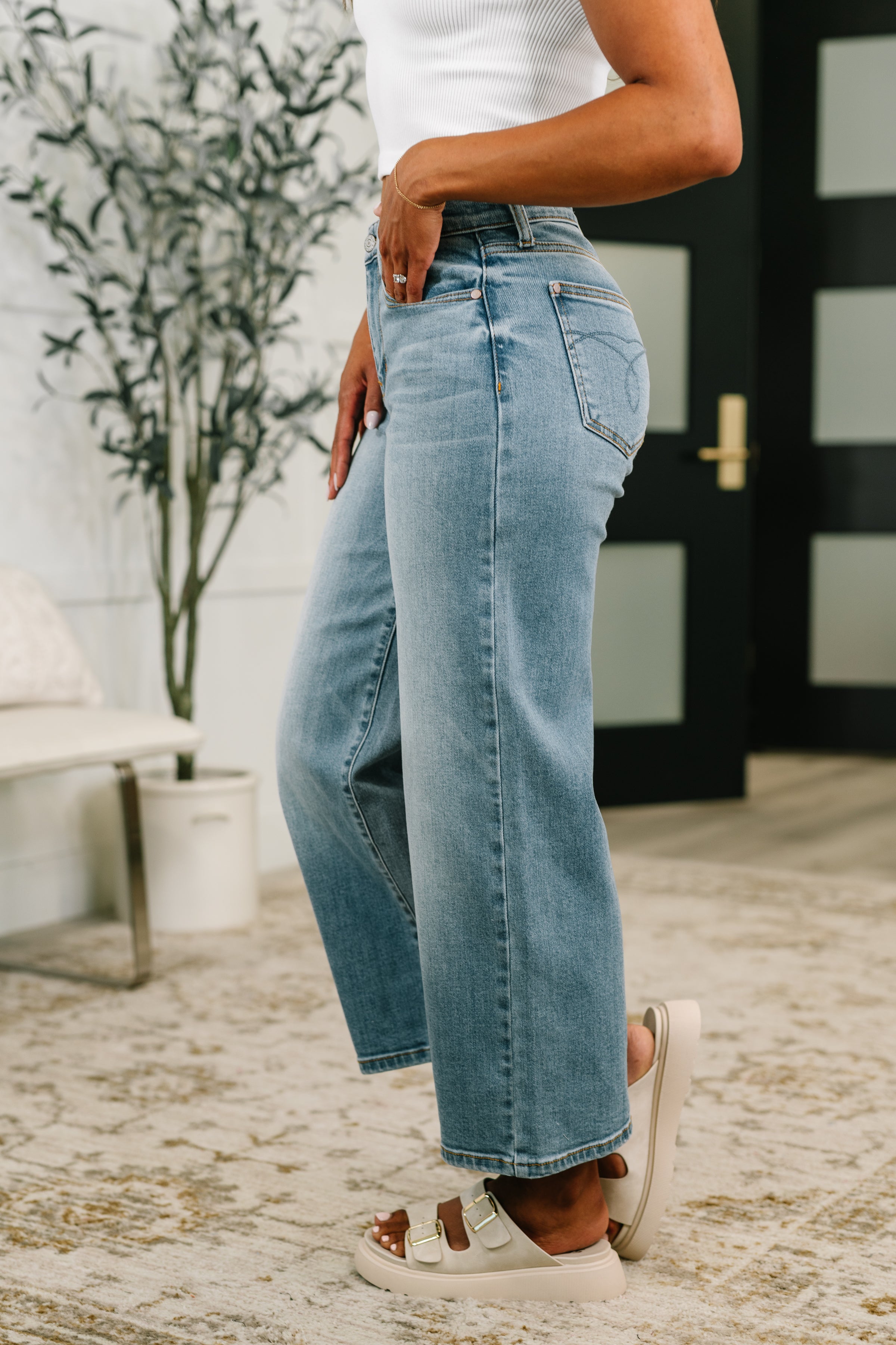 Side view of light blue mid-rise wide leg ankle jeans paired with white buckle sandals indoors