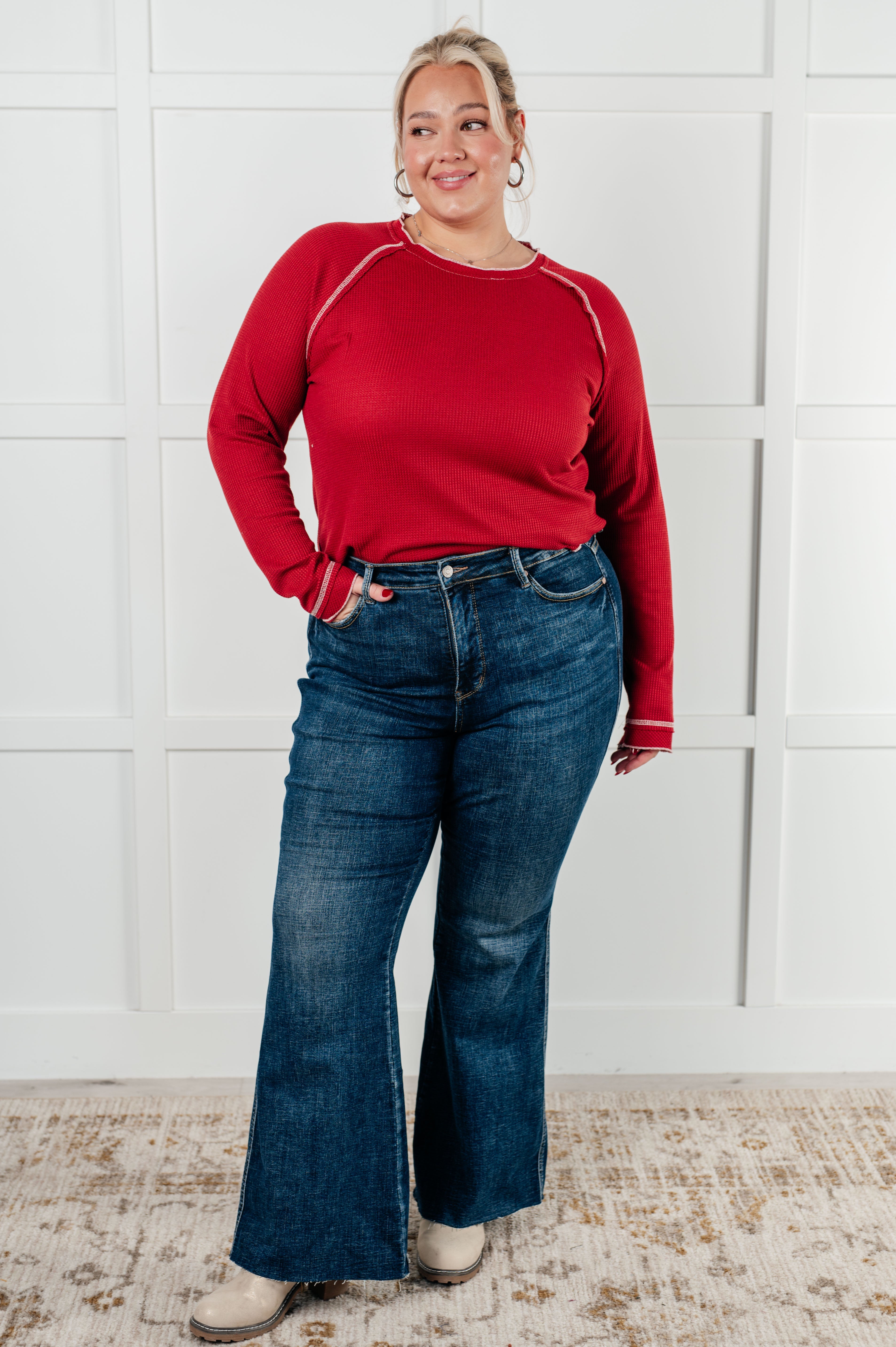 Woman modeling red Clever Expression Long Sleeve Top with contrast stitching paired with dark flared jeans and boots indoors
