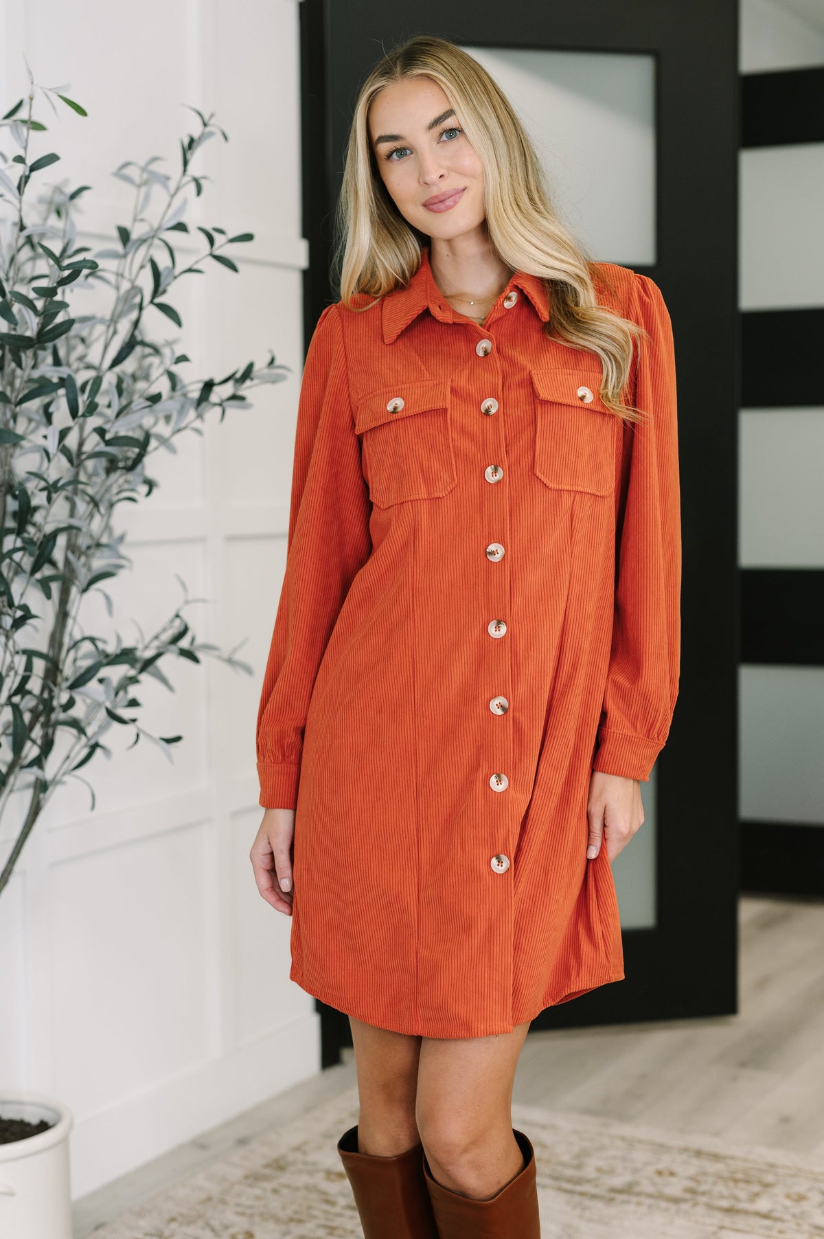 Woman modeling a rust corduroy shirt dress with a collared neckline front buttons patch pockets and gathered shoulders with tall brown boots indoors