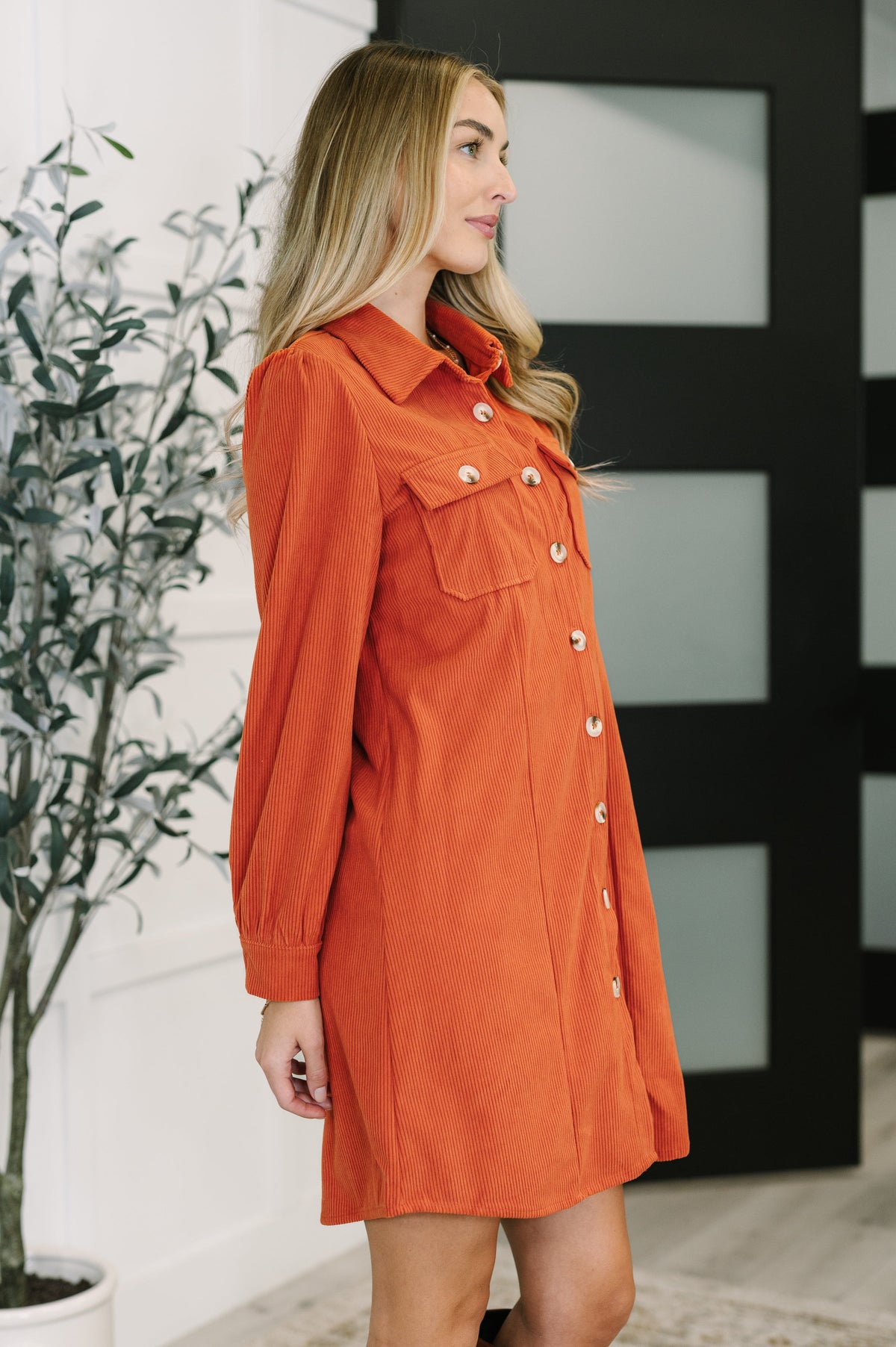 Woman modeling a rust corduroy shirt dress with a collared neckline button front patch pockets and gathered shoulders indoors