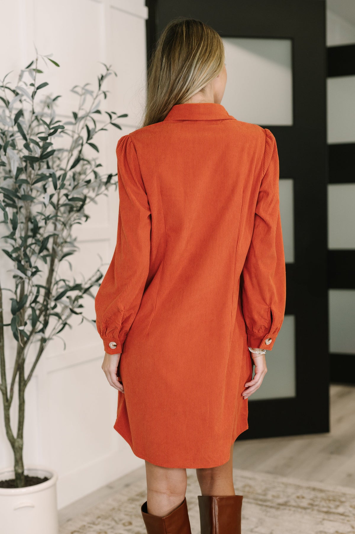Back view of a woman wearing a rust corduroy shirt dress with gathered shoulders and long sleeves in a modern indoor setting
