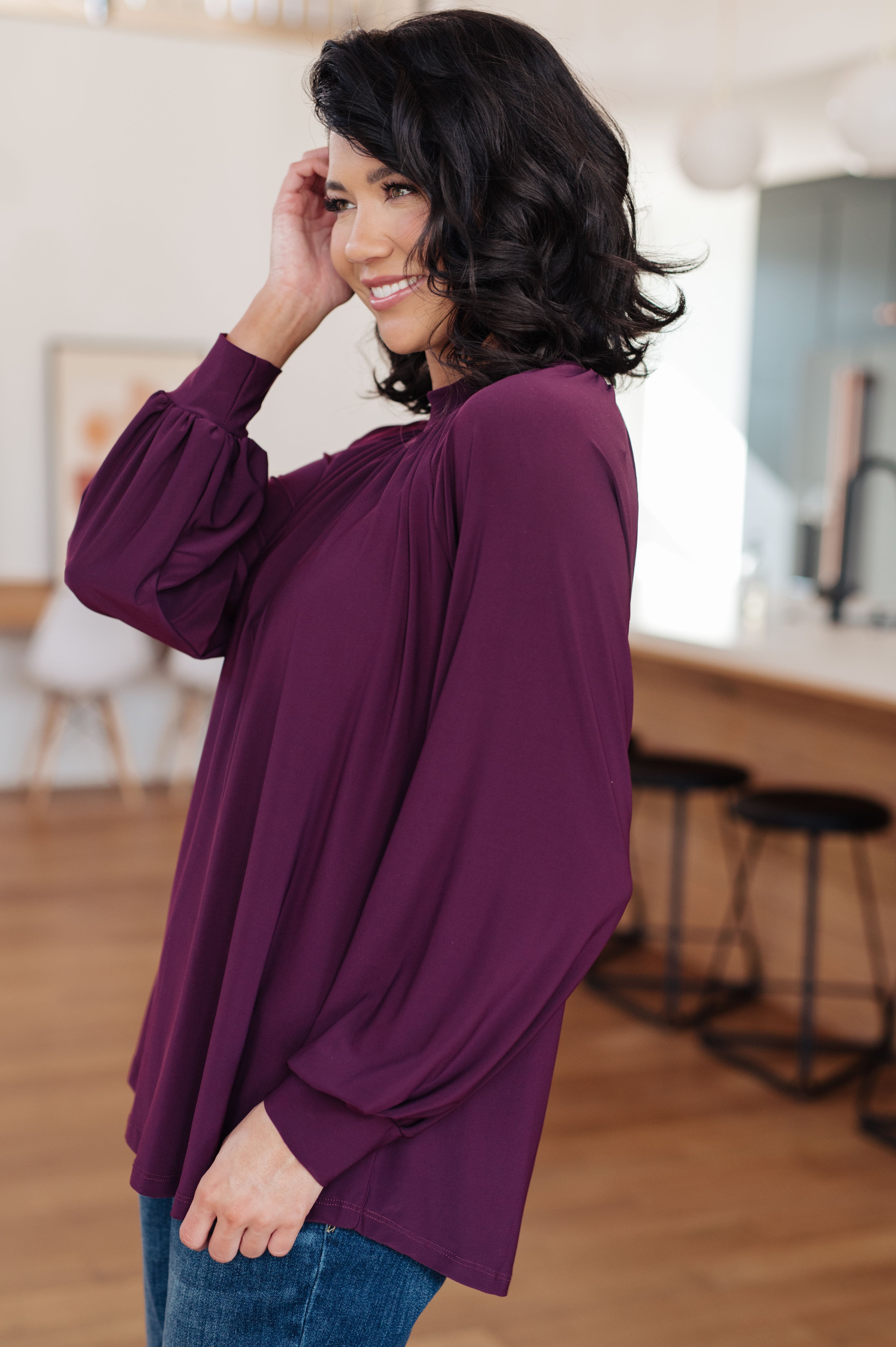 Side view of woman smiling and wearing a plum-colored mock neck blouse with gathered sleeves indoors