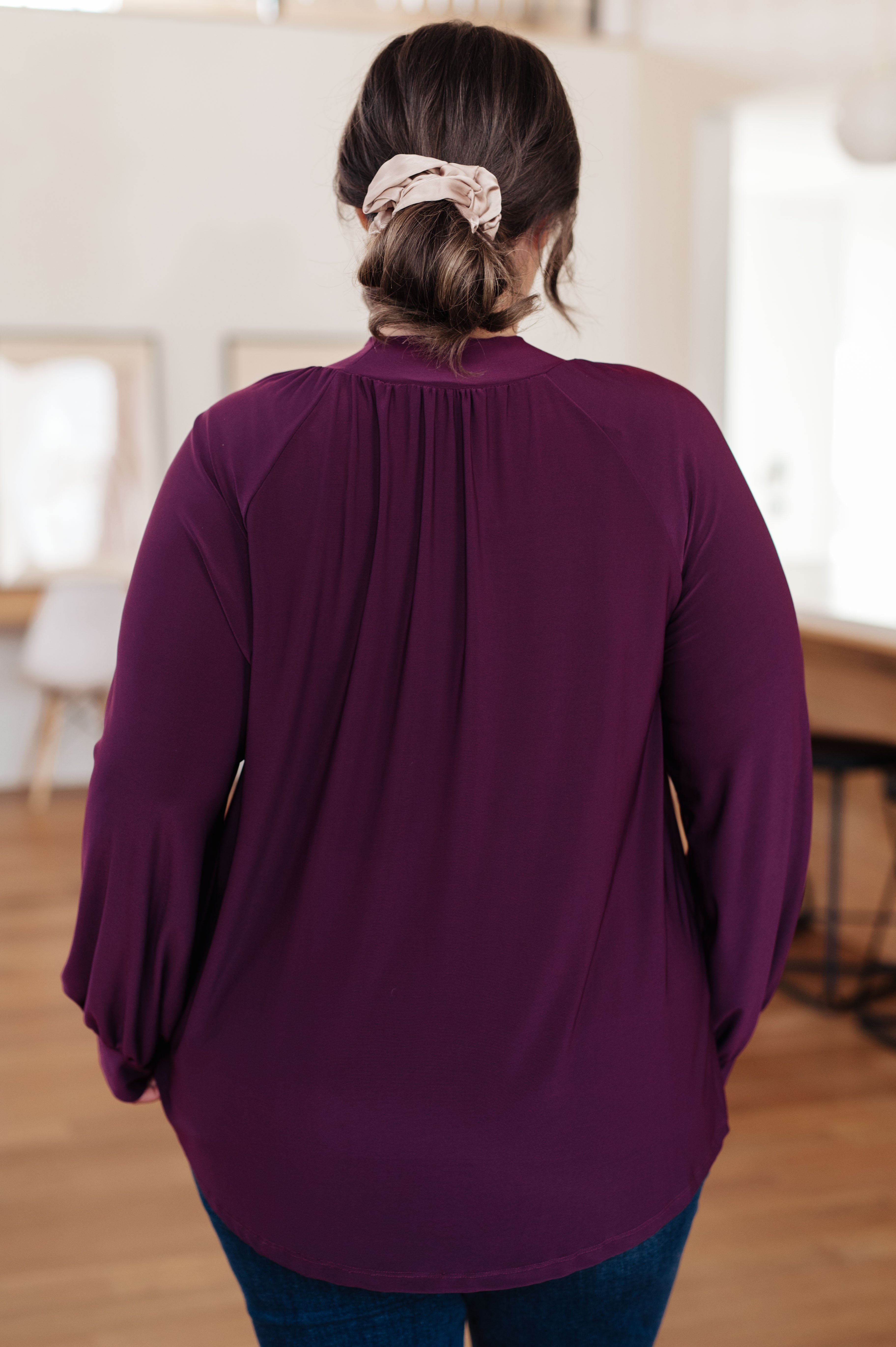 Back view of woman wearing deep purple mock neck blouse with gathered yoke and long sleeves in indoor setting