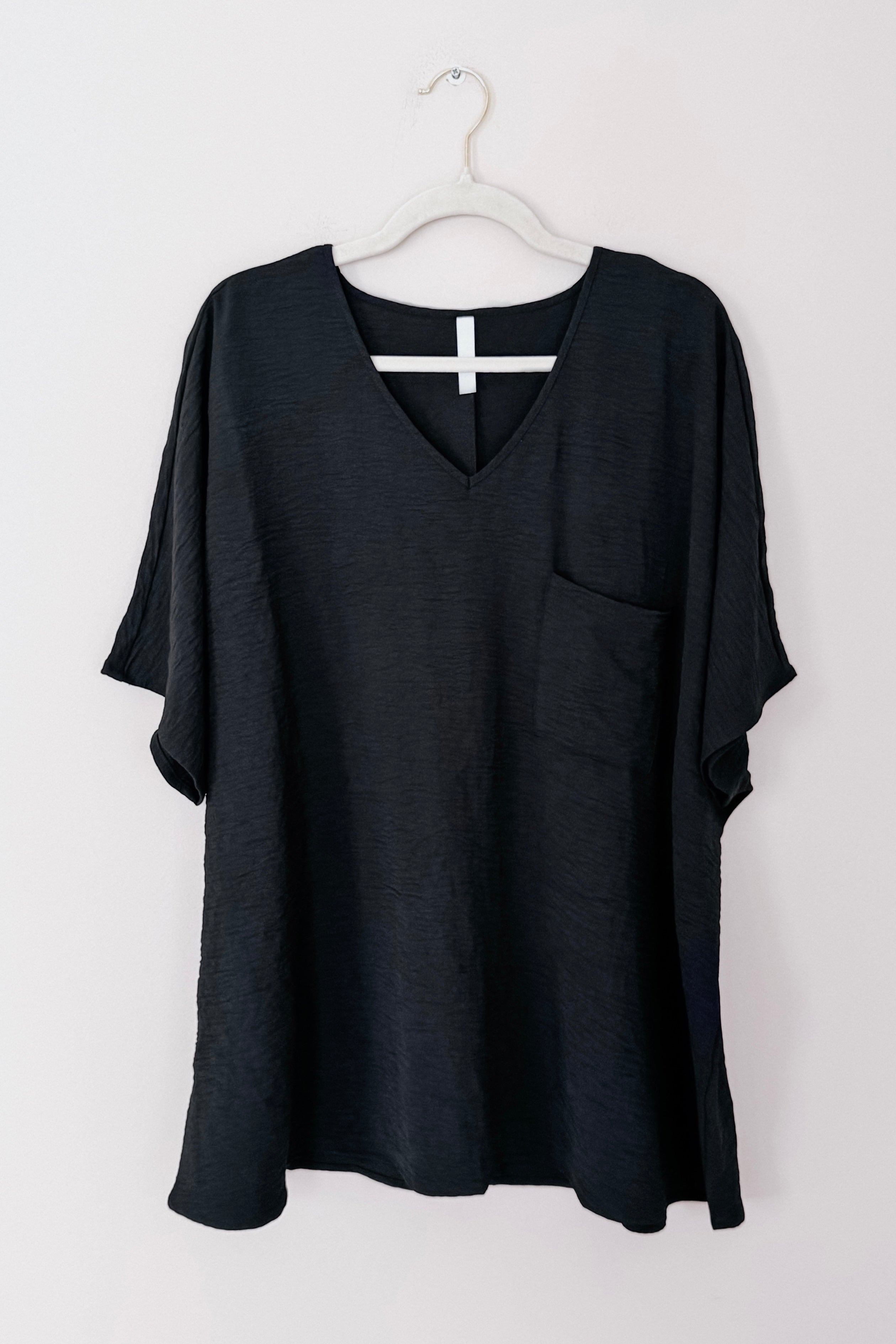 Black loose-fit V-neck top with short sleeves and front pocket hanging on white hanger against light background
