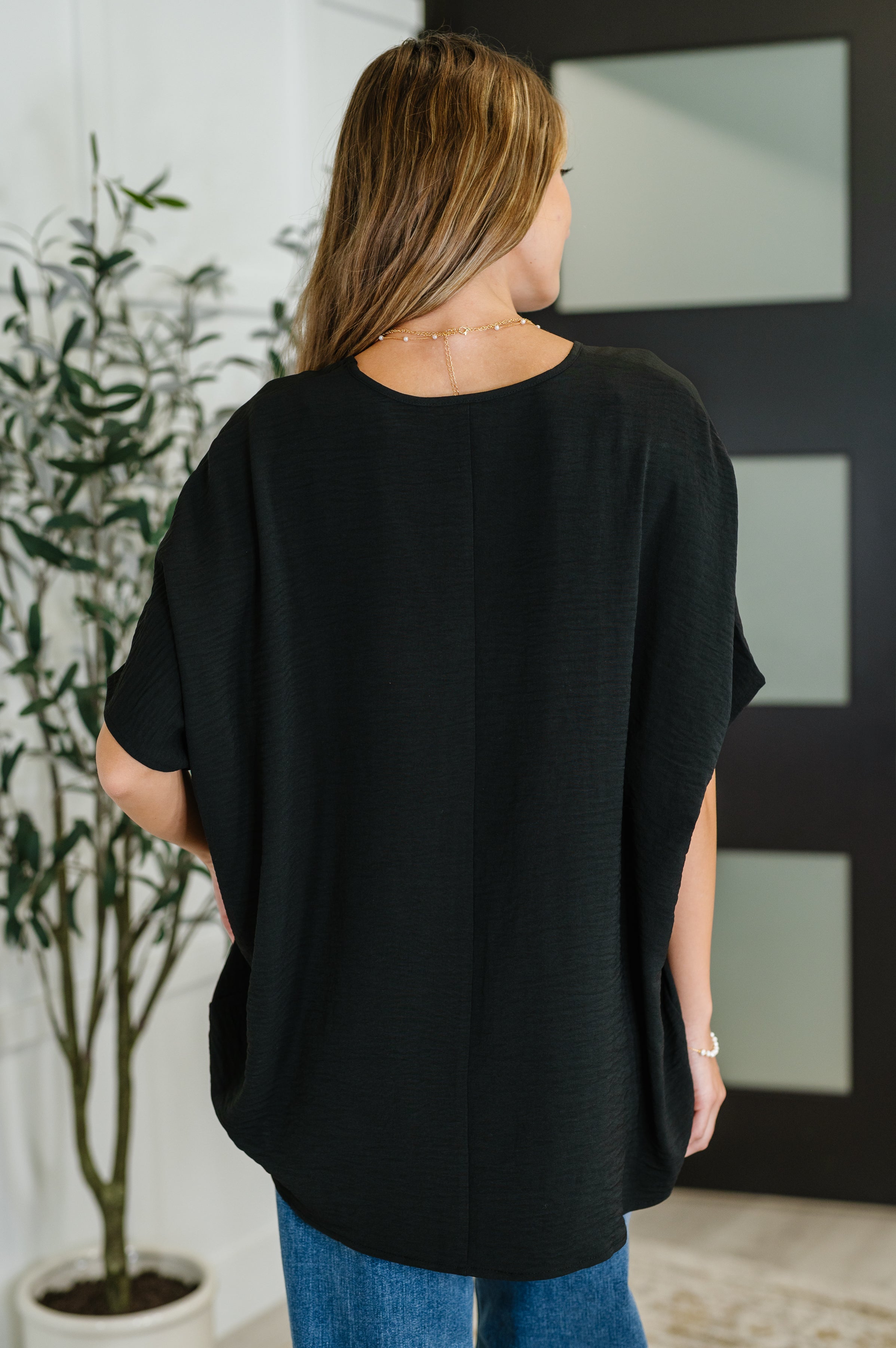Back view of woman wearing loose black V-neck top with short sleeves standing indoors near plant and door