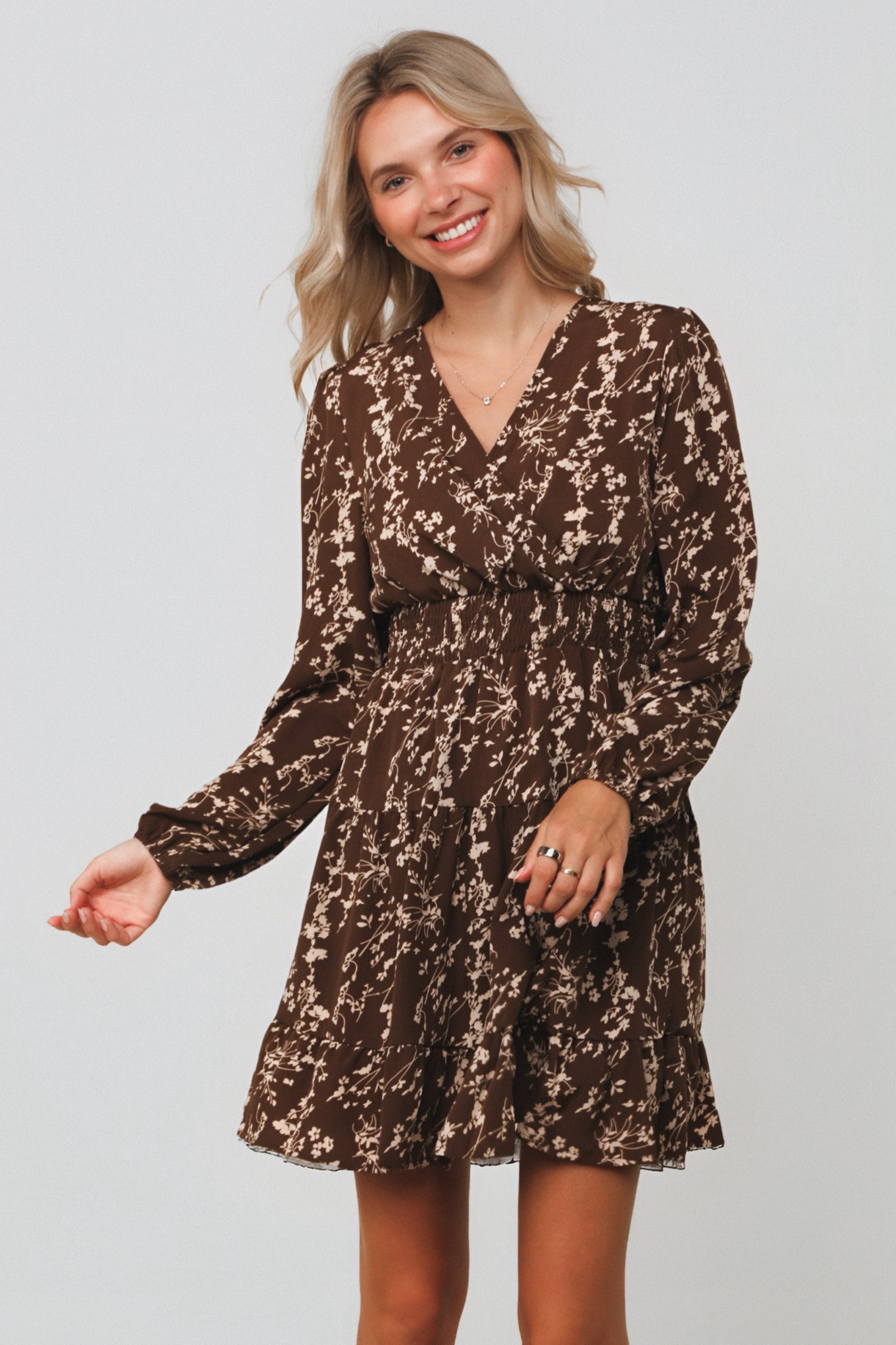 Smiling woman wearing brown floral smocked waist dress with long sleeves and V-neck standing against plain background