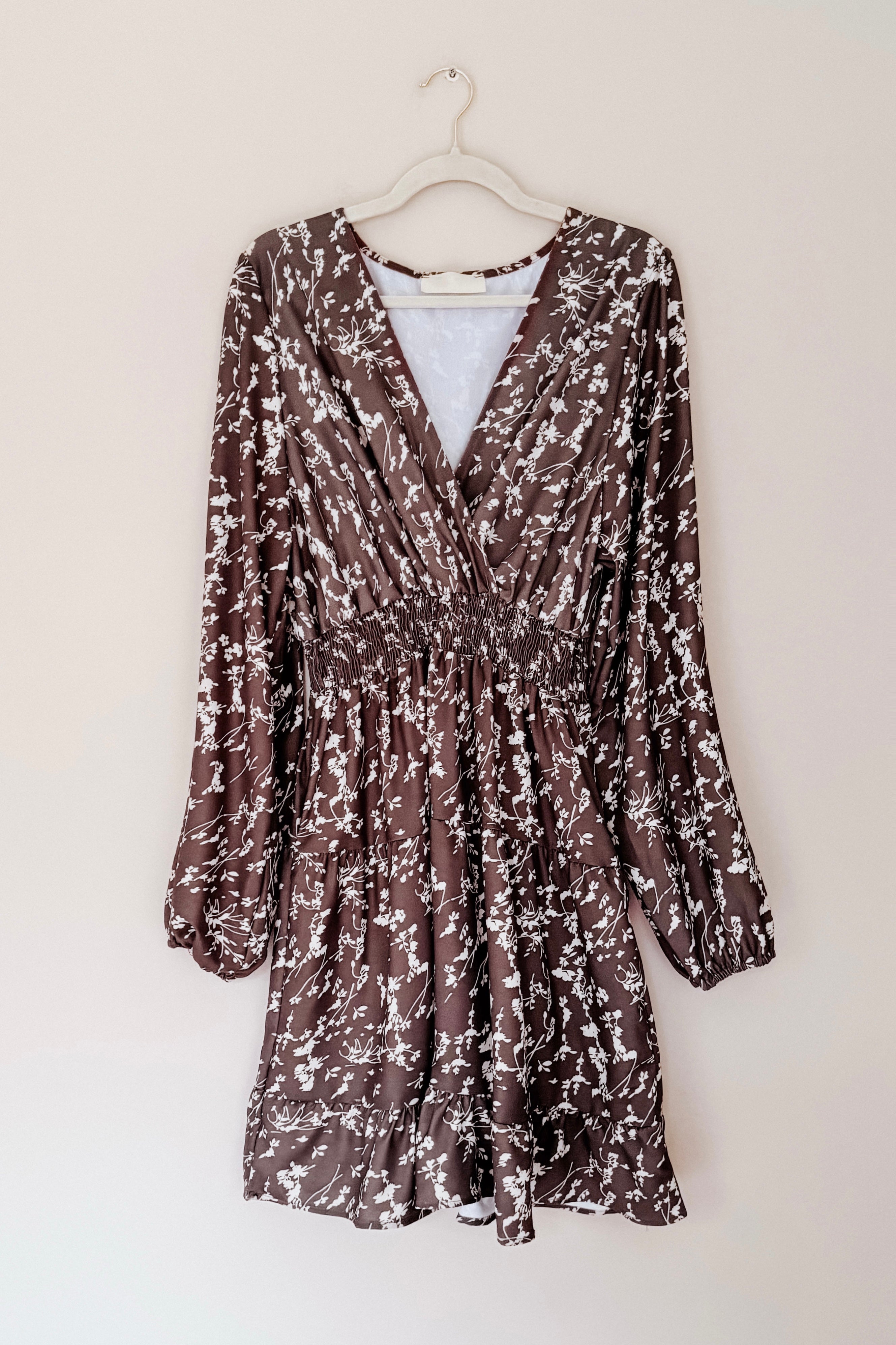 Brown floral print dress with smocked waist, long sleeves, and V-neck hanging on white hanger against beige wall