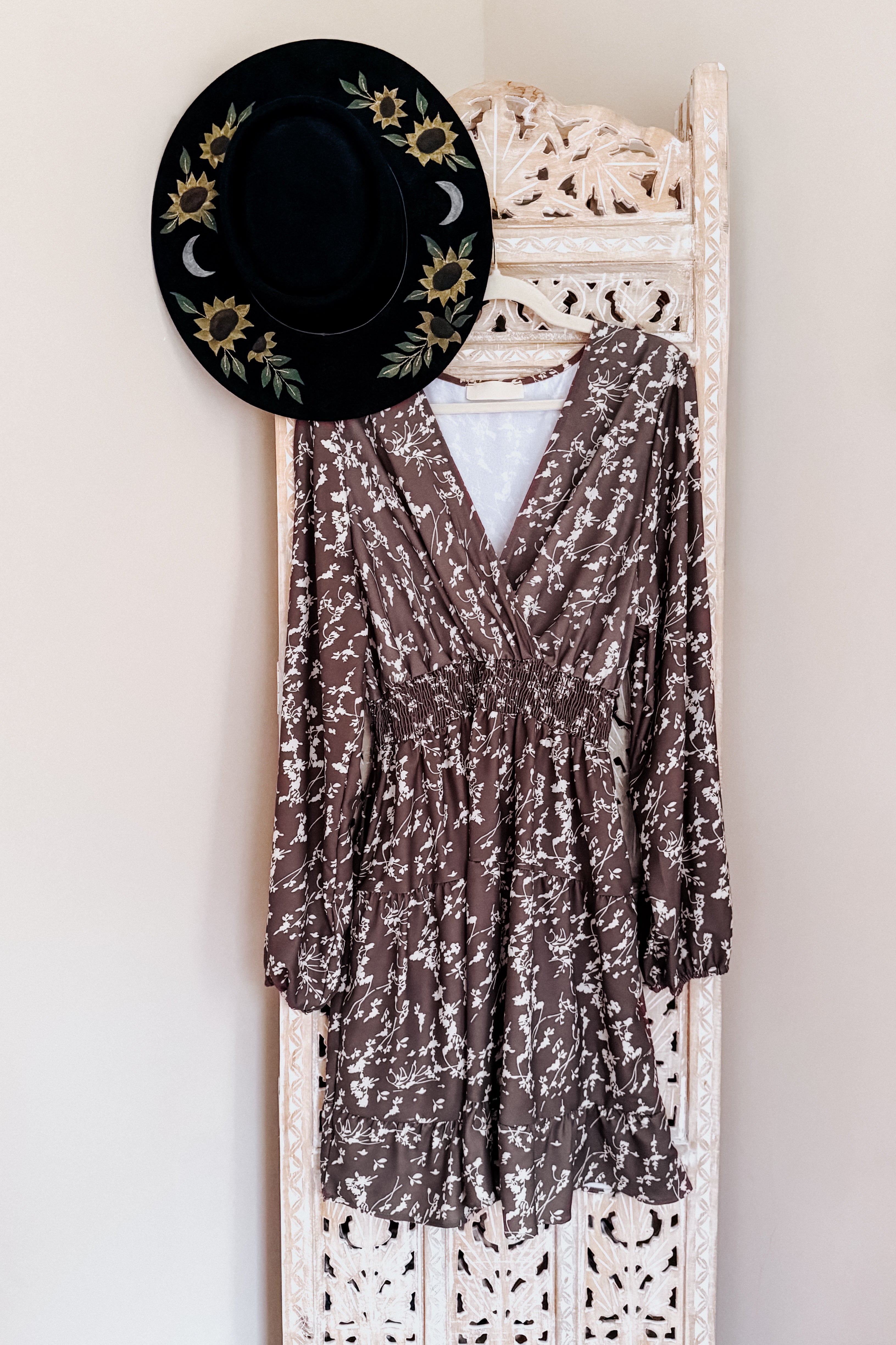 Cocoa brown floral wrap dress with smocked waist and long sleeves hanging on carved wooden screen with black sunflower hat