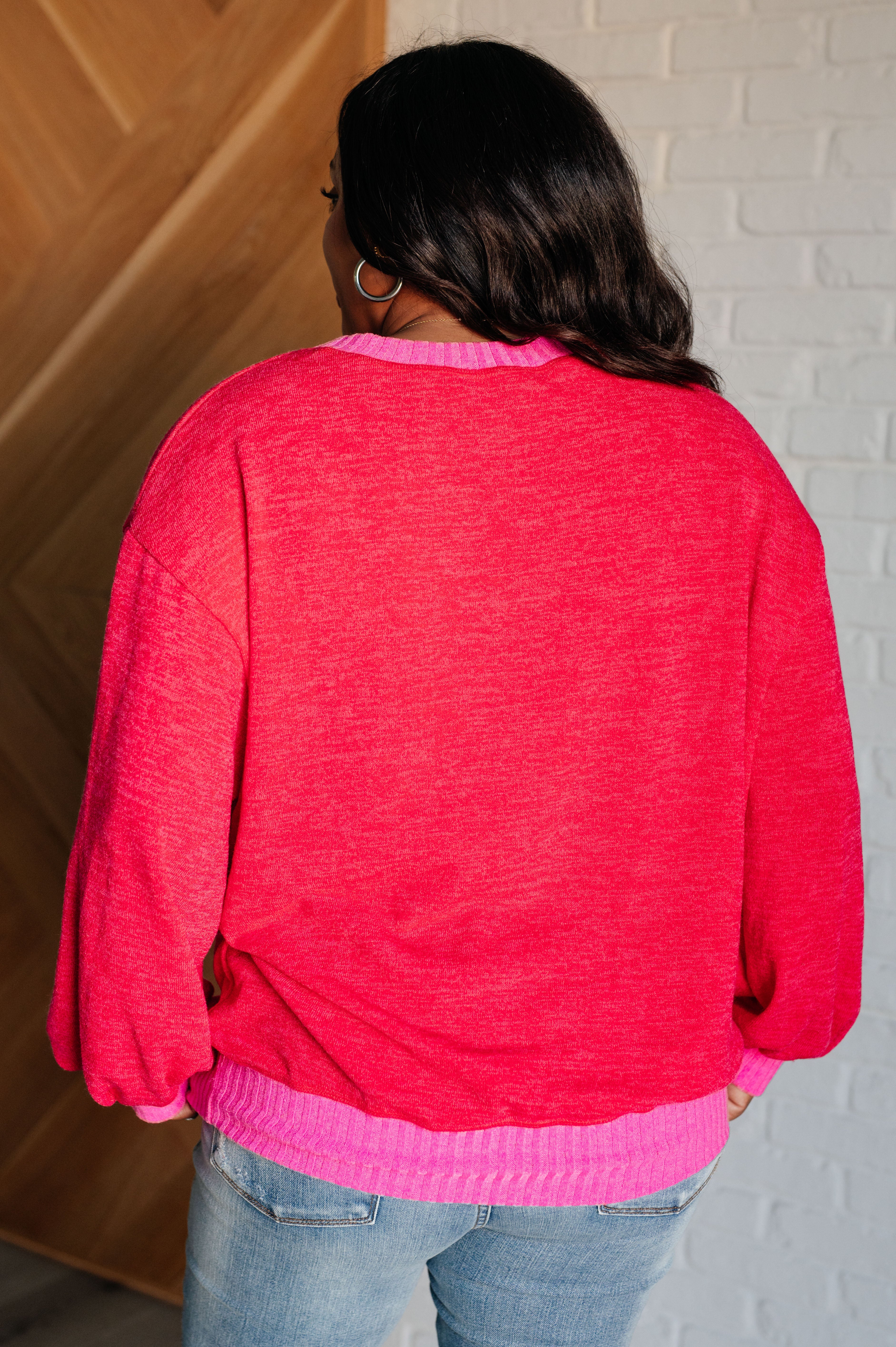 Woman wearing a red sweater with bright pink trim and blue jeans standing against a white brick wall background