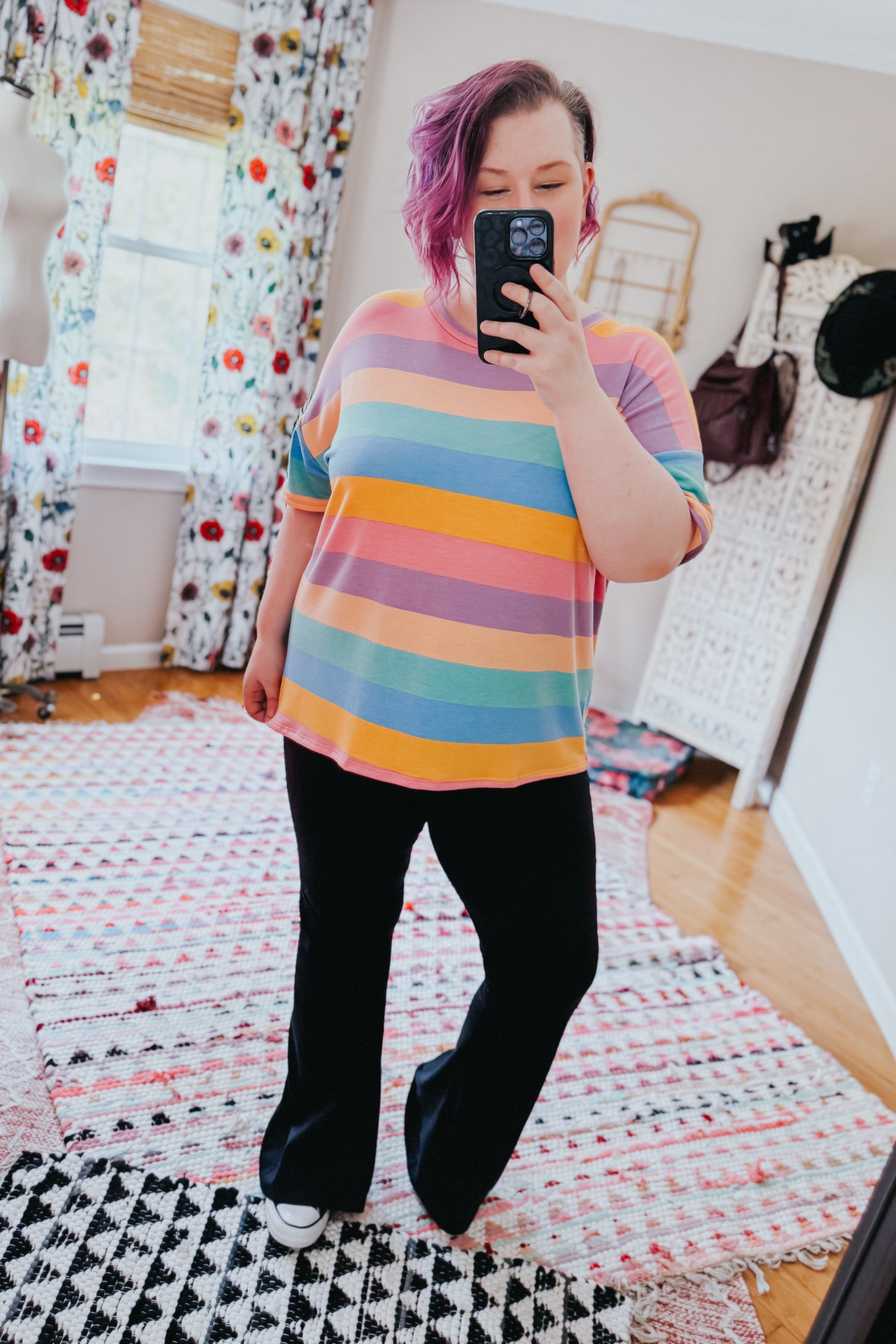 Woman wearing pastel rainbow striped drop shoulder top with black pants taking mirror selfie indoors