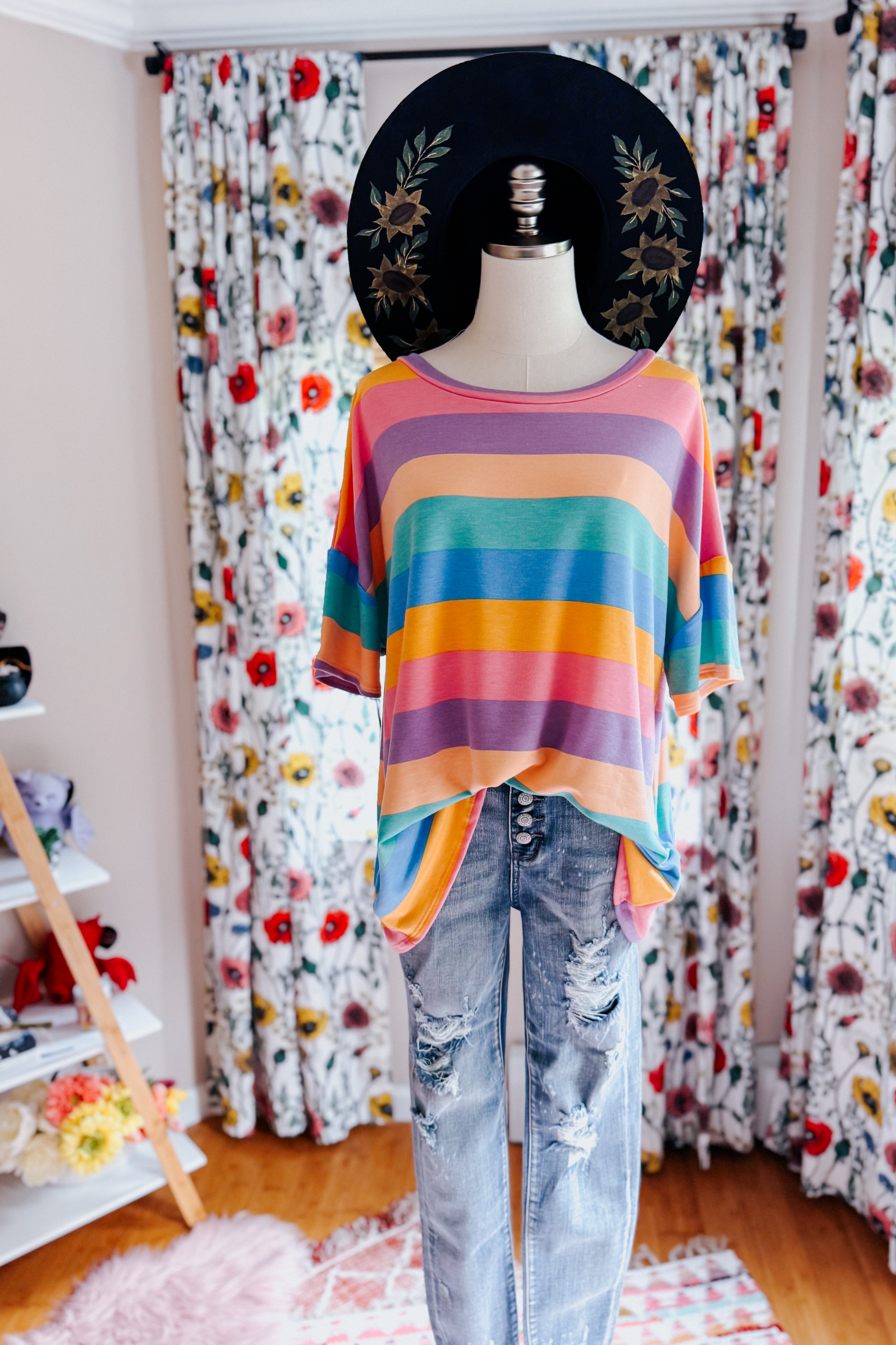 Mannequin dressed in colorful horizontal striped drop shoulder top paired with distressed light wash jeans and black floral hat