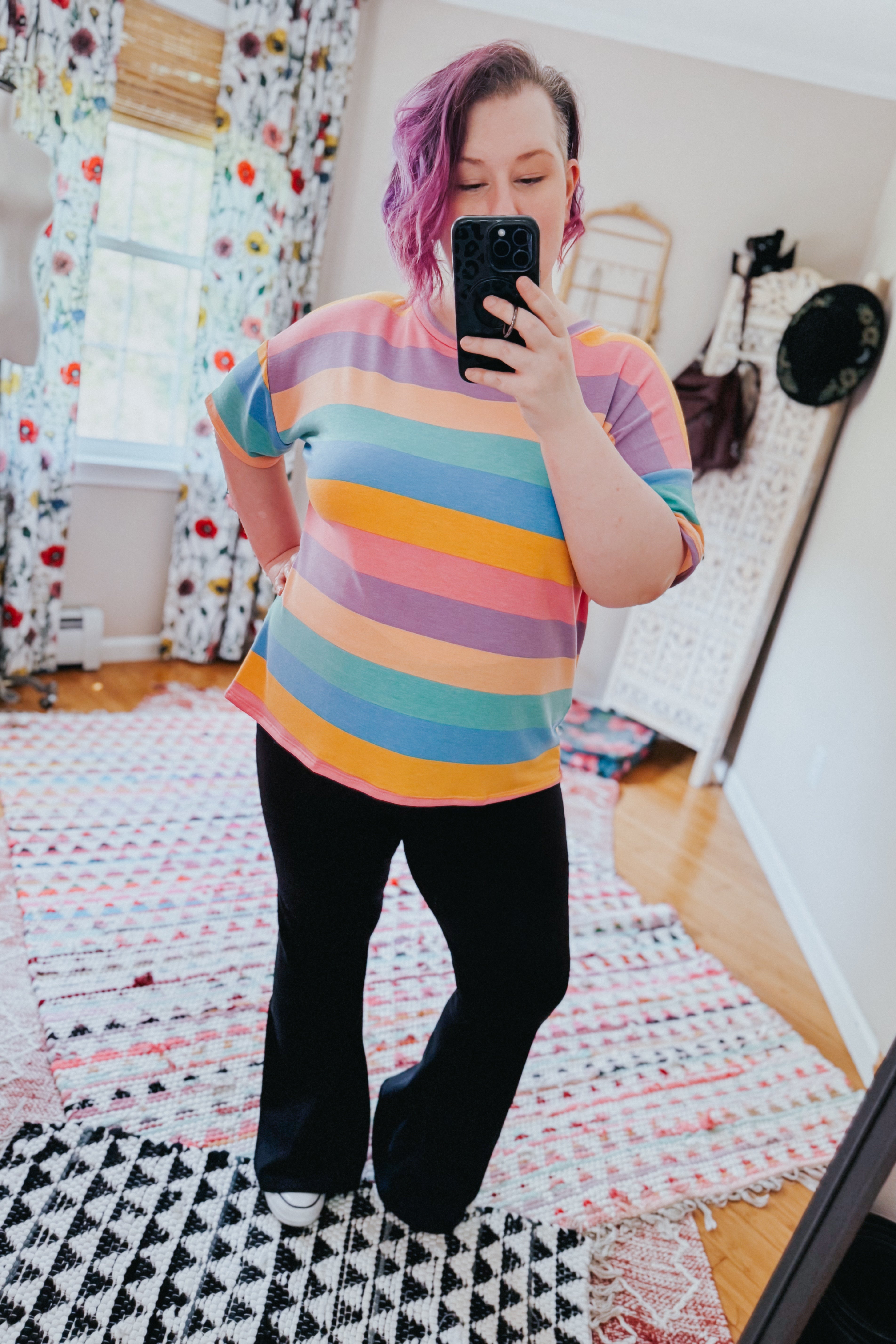 Woman taking mirror selfie wearing pastel rainbow striped drop shoulder top and black pants in bright room