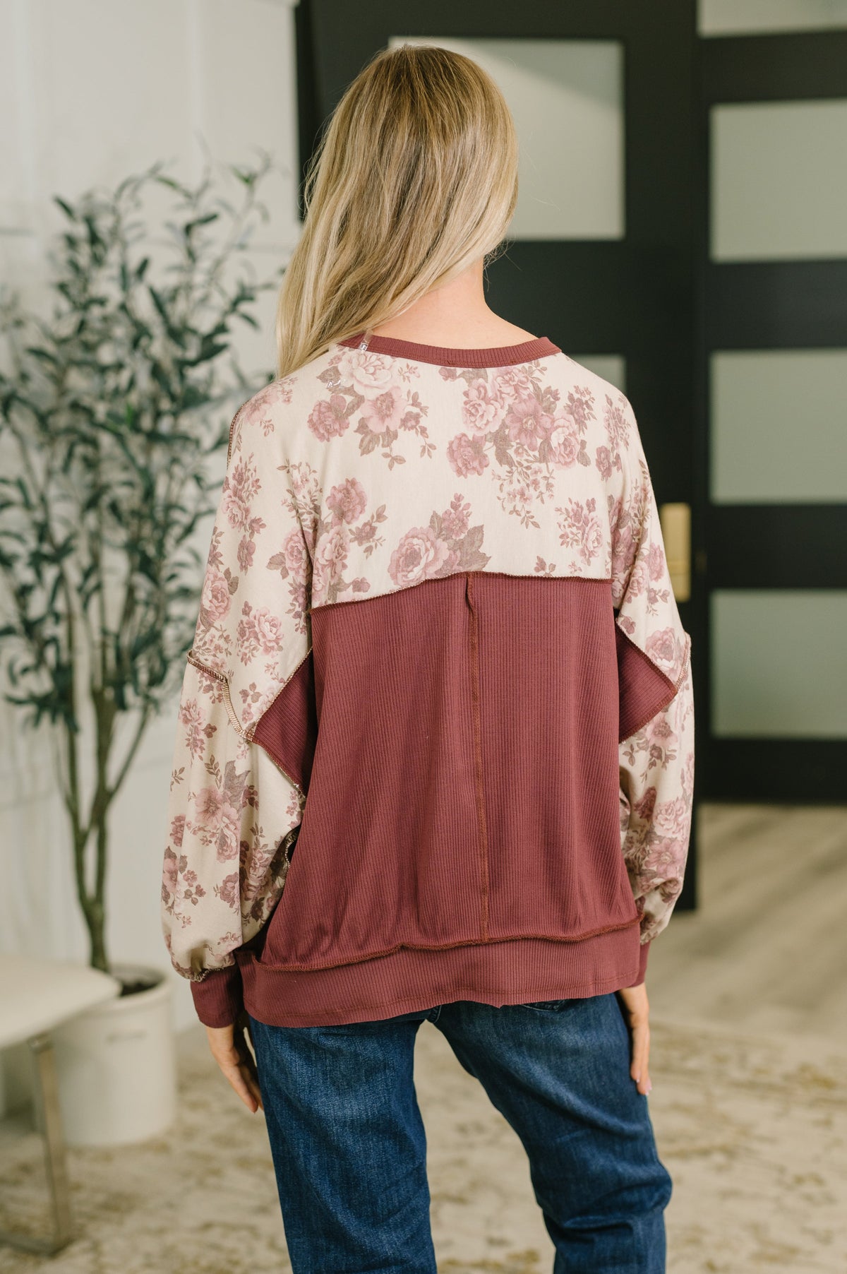 Back view of a woman wearing a rust pullover with a floral print yoke and sleeves styled with blue jeans indoors