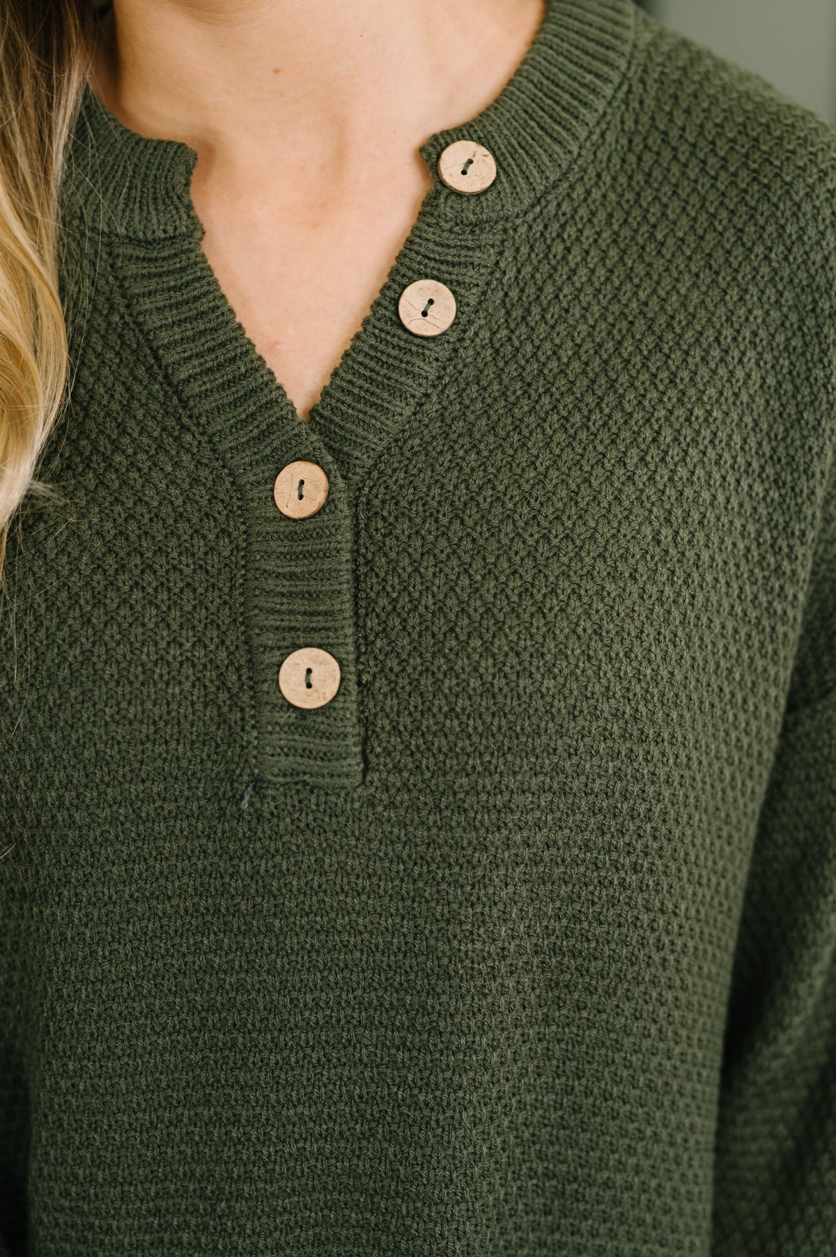 Close up of a dark olive knit henley top featuring a V neckline and four natural tone buttons on the front placket