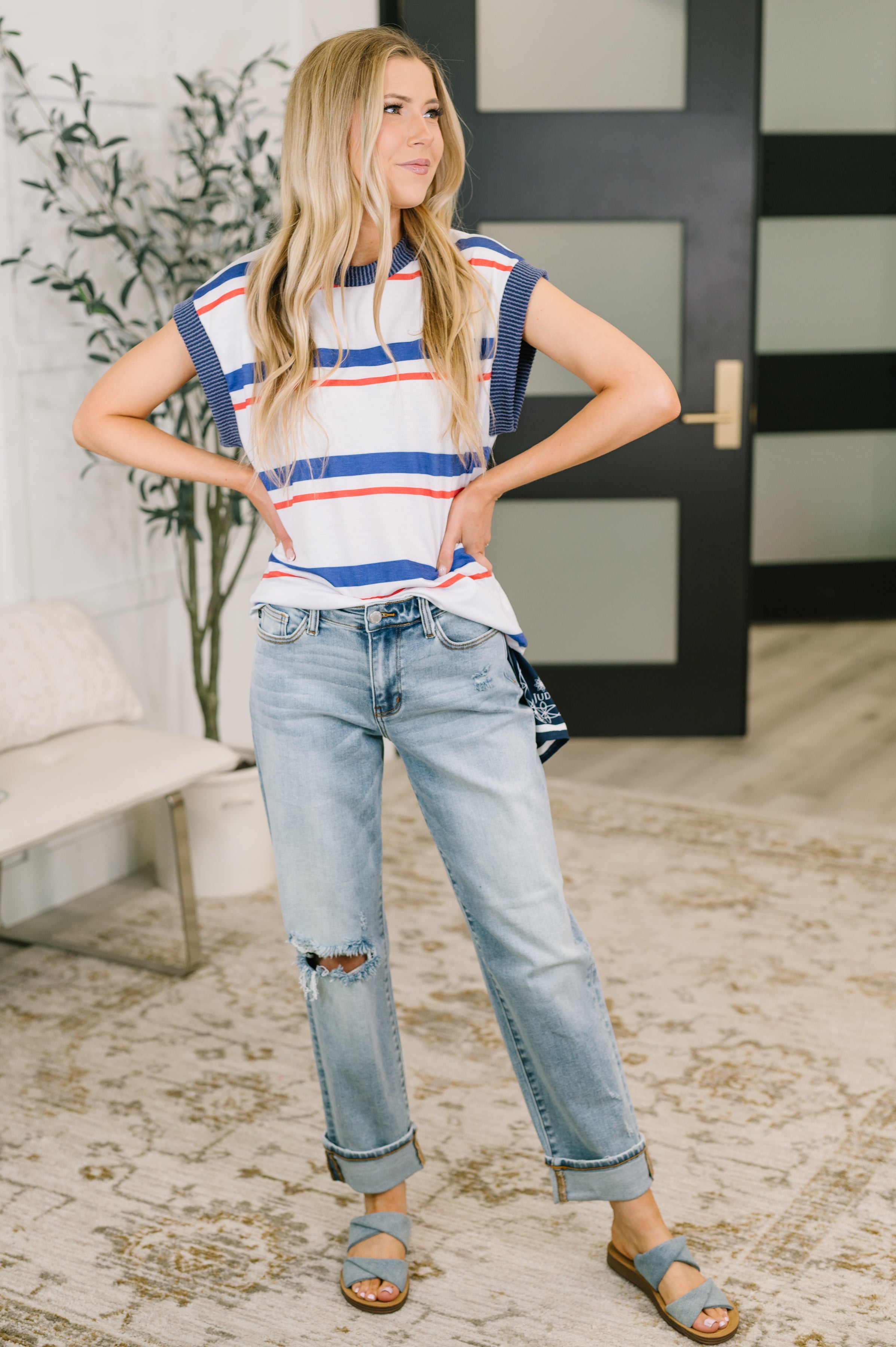 Blonde woman wearing a blue and red striped sleeveless top paired with light distressed denim jeans and sandals indoors