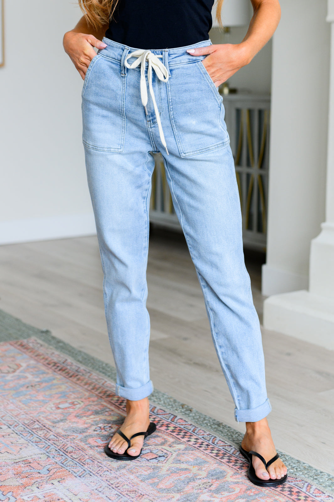 Woman wearing light wash high rise denim joggers with front patch pockets and drawstring tie standing indoors