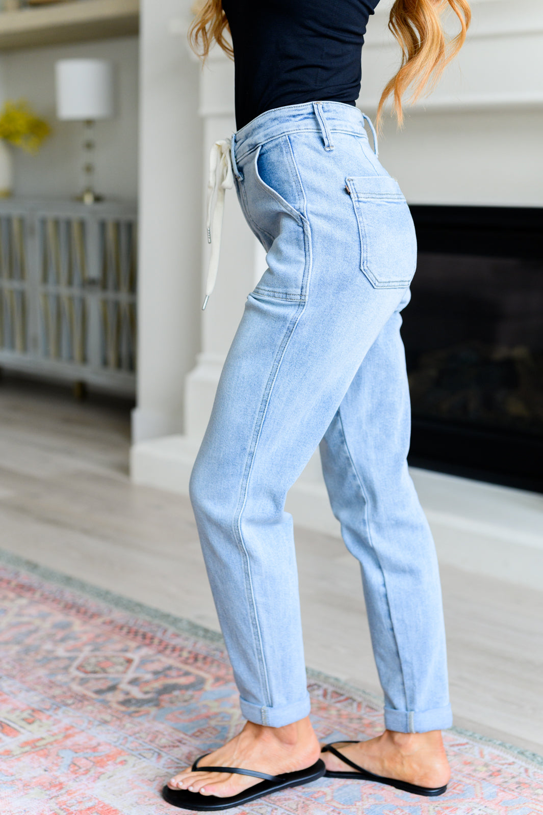 Woman wearing light wash high rise denim joggers with cuffed hems standing indoors on patterned rug near fireplace