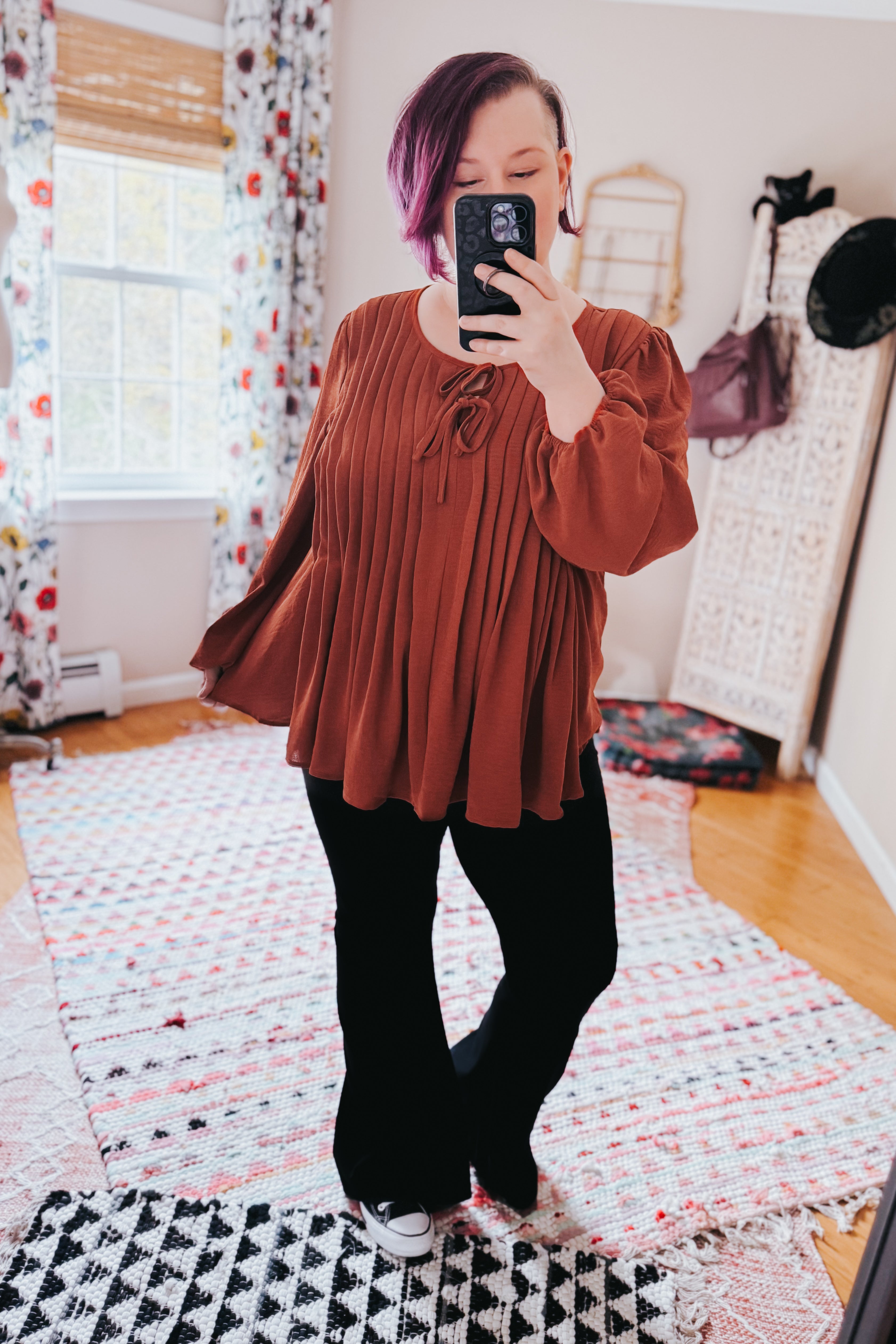 Woman taking mirror selfie wearing flowing copper pleated blouse with long sleeves and tie neckline with black pants indoors