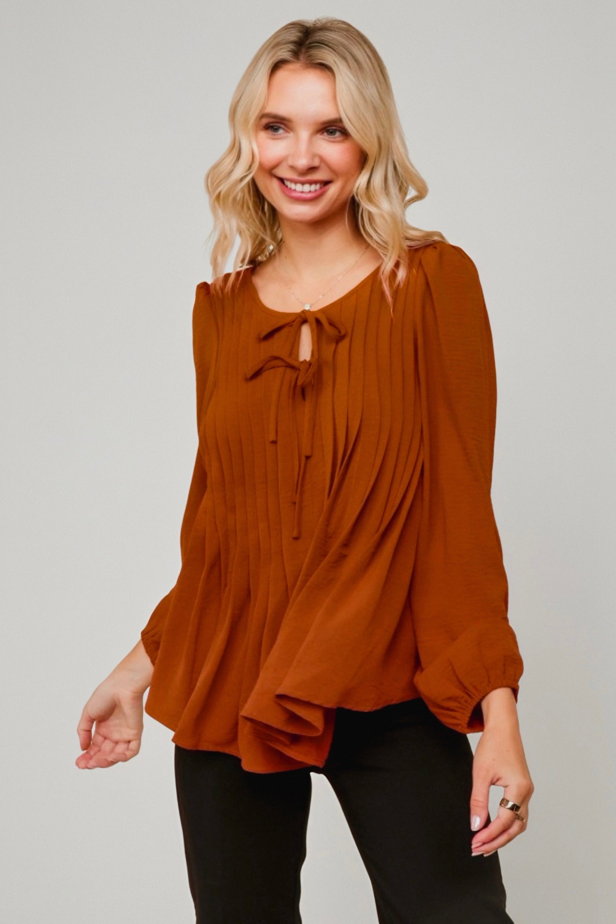 Smiling woman wearing flowing copper pleated blouse with balloon sleeves and tie neckline paired with black pants