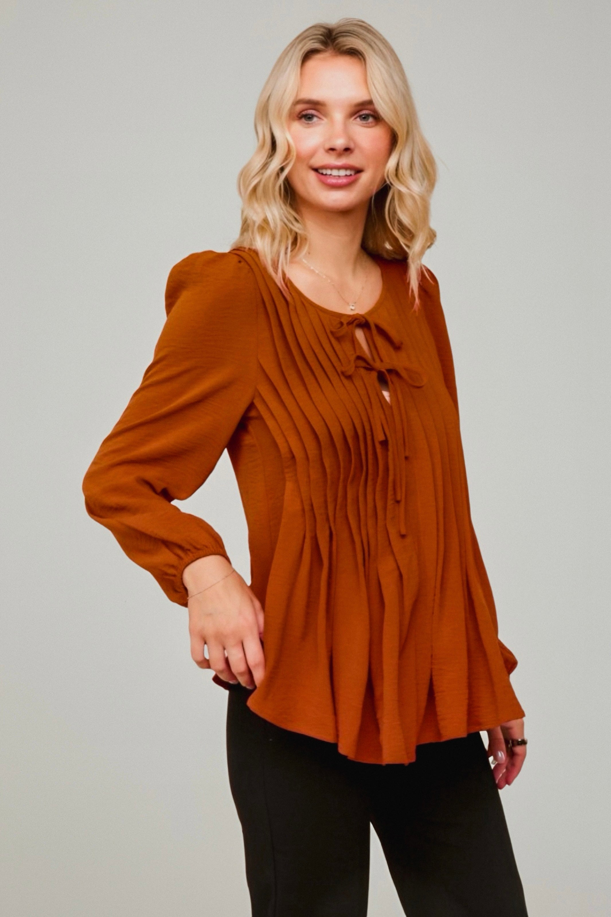 Blonde woman wearing flowy copper pleated blouse with long sleeves and front tie detail paired with black pants against grey background