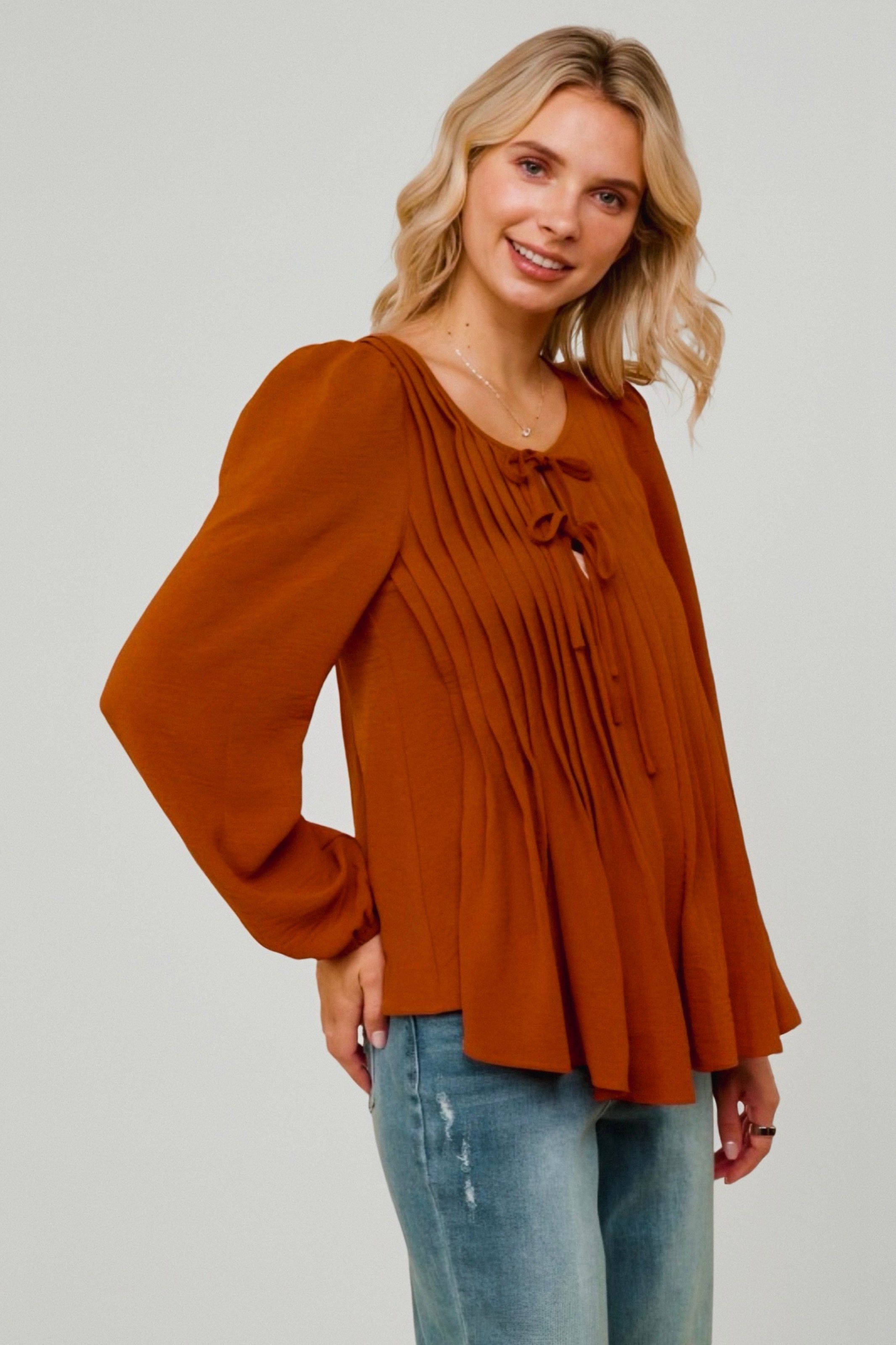 Woman wearing burnt orange pleated blouse with long puff sleeves and tie detail at neckline paired with distressed jeans
