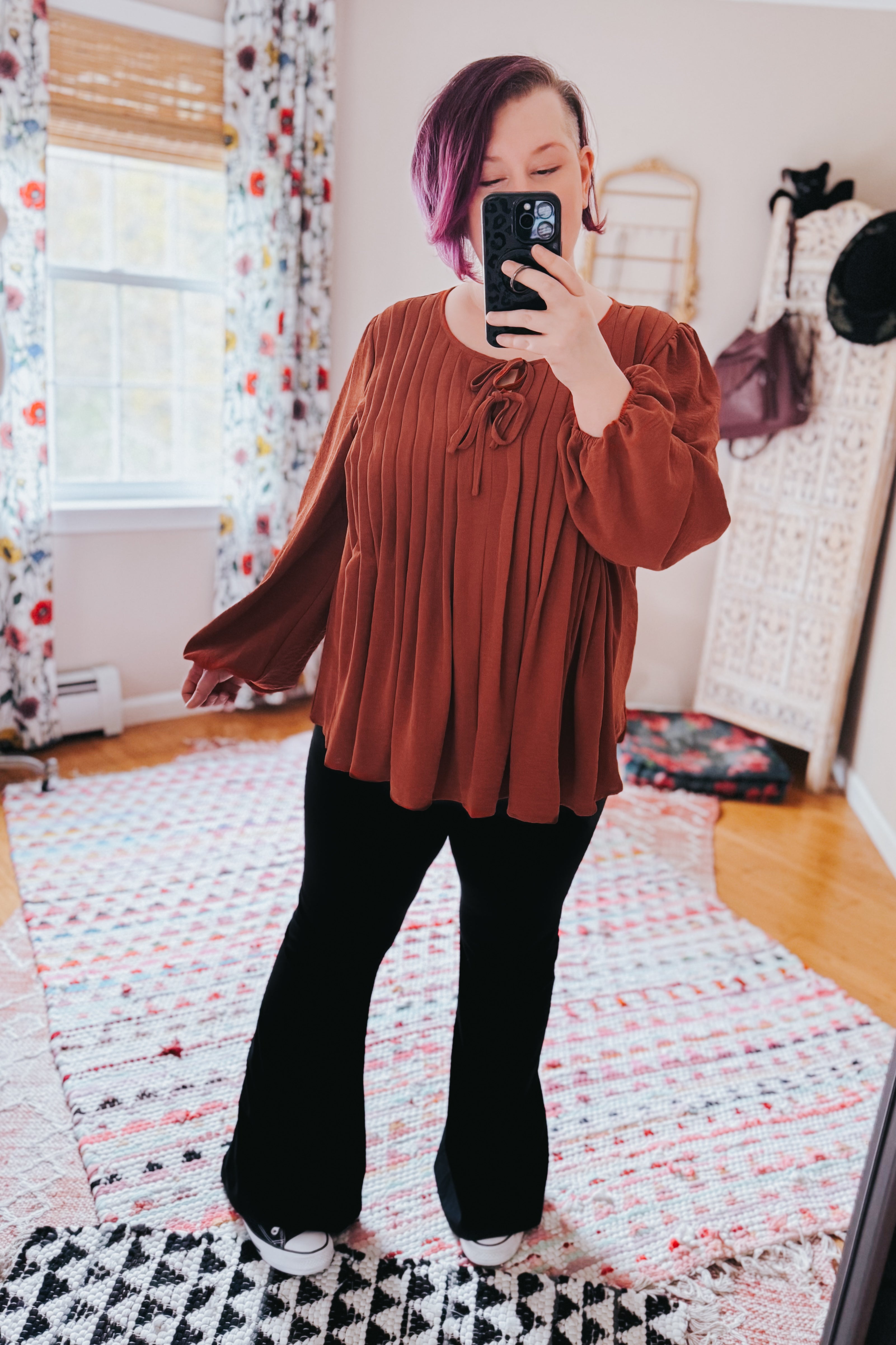Woman posing indoors wearing flowing copper pleated blouse with long bishop sleeves and tie detail at neckline with black pants