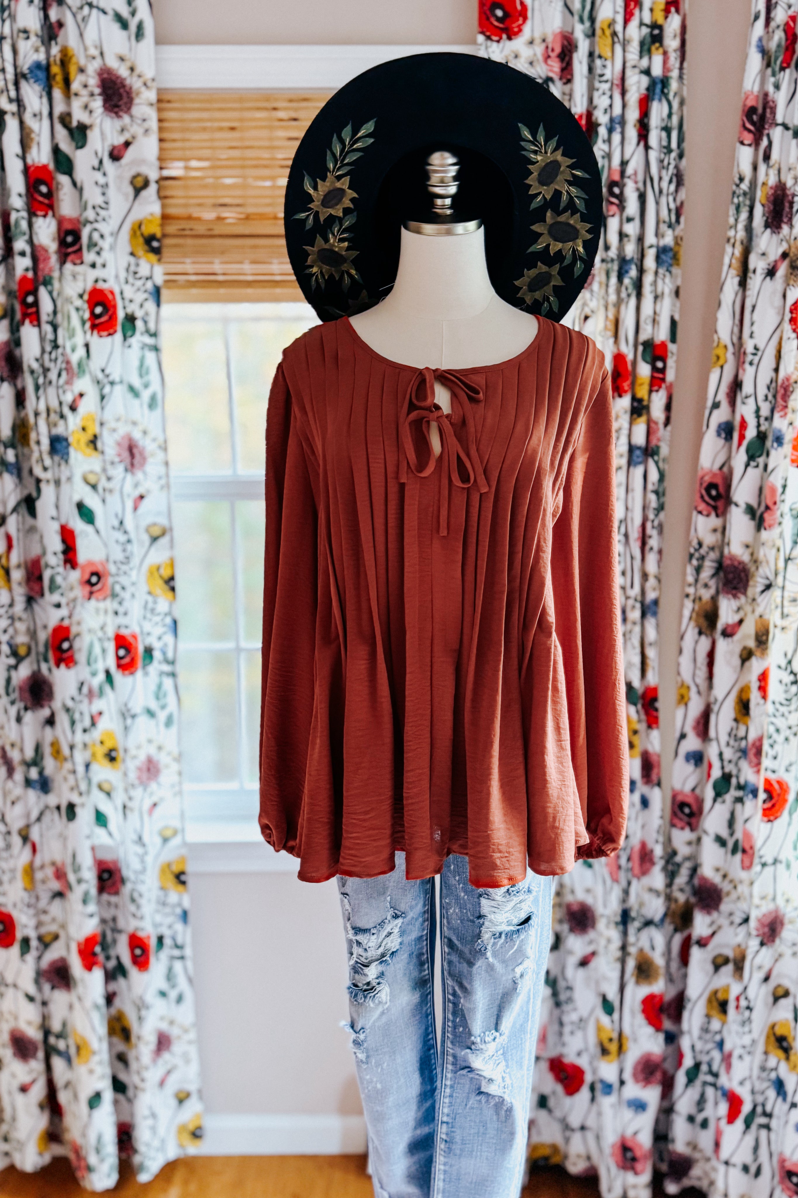 Rust-colored pleated blouse with long sleeves and double tie neckline styled on mannequin with floral black wide-brimmed hat