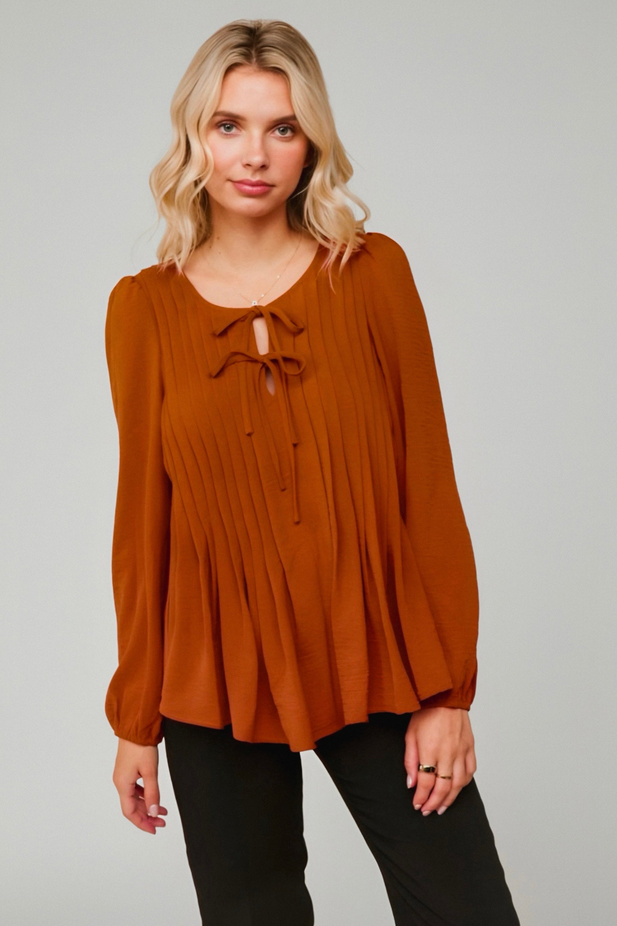 Woman wearing copper pleated blouse with long sleeves and double tie detail at neckline paired with black pants against grey background