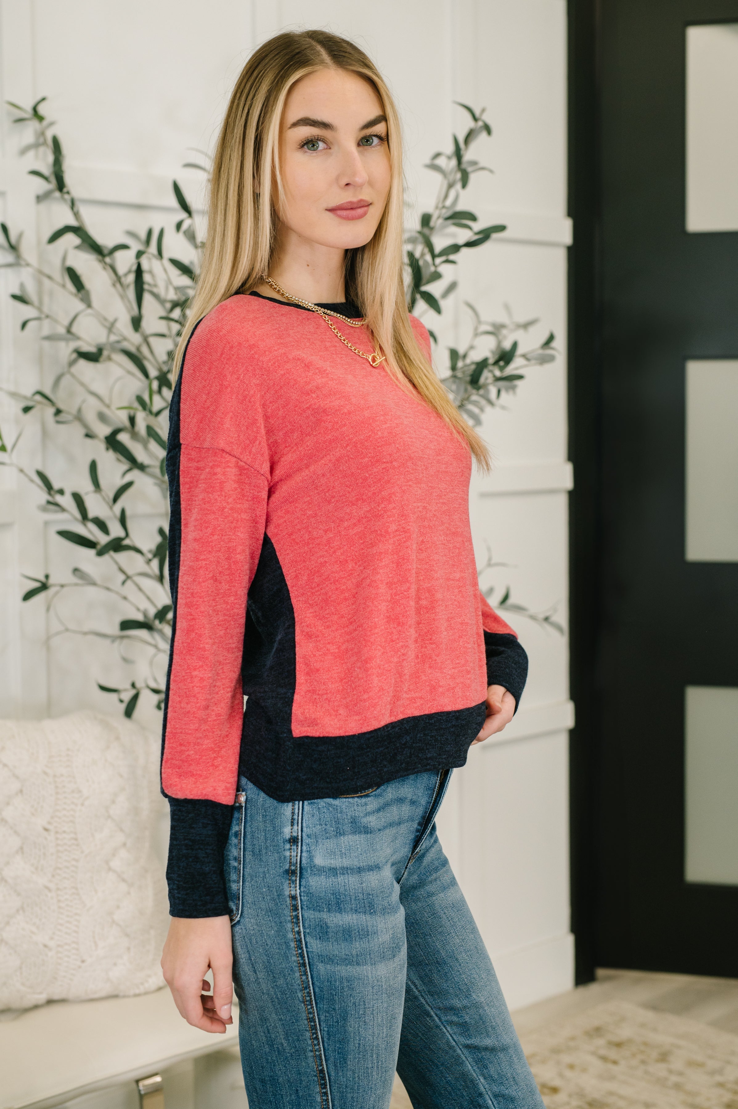 Woman wearing a red sweater with black trim and blue jeans standing indoors with decorative plants in the background.