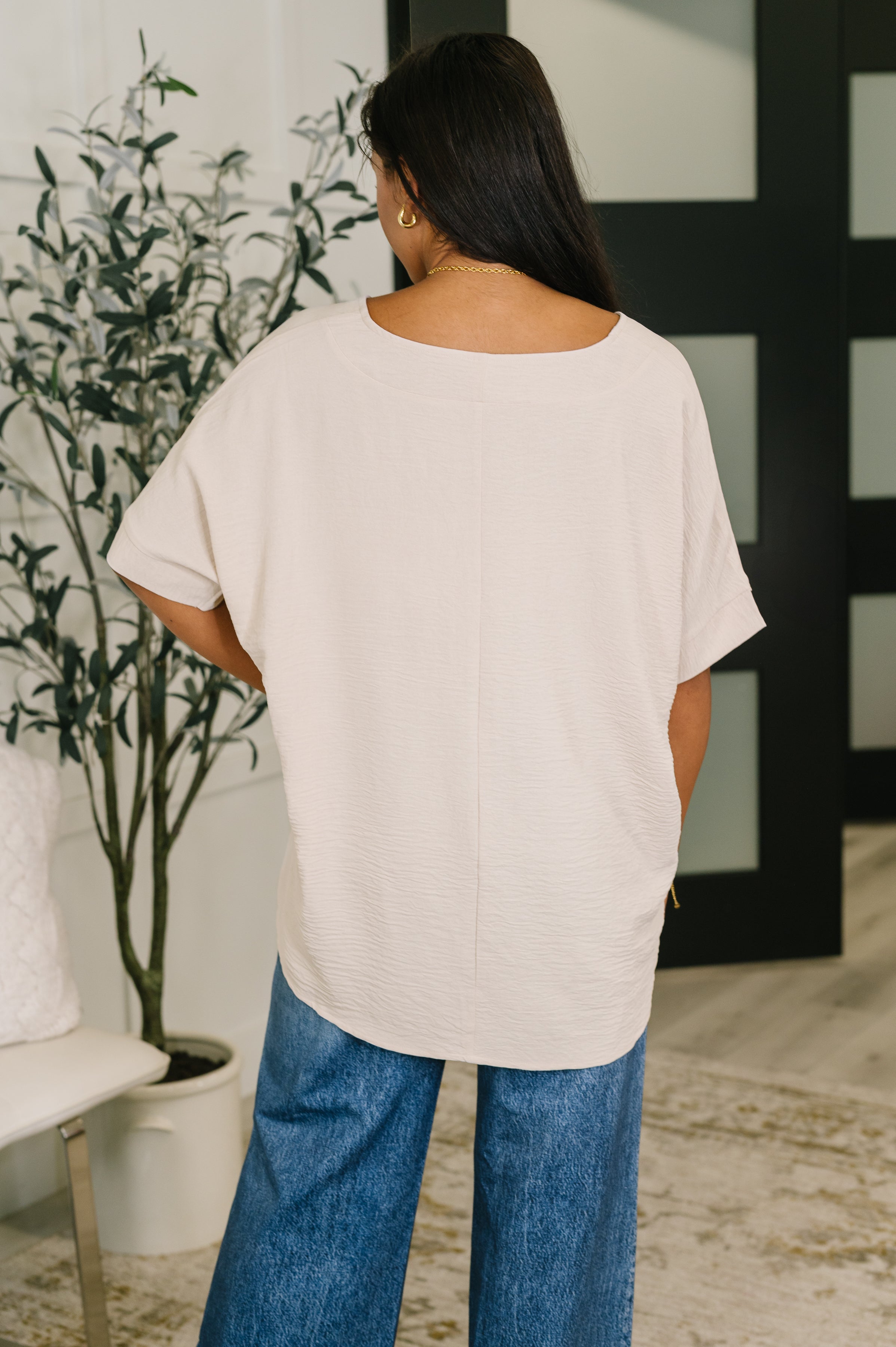 Woman wearing a sand beige crinkle textured relaxed v-neck top with dolman sleeves in an eclectic room