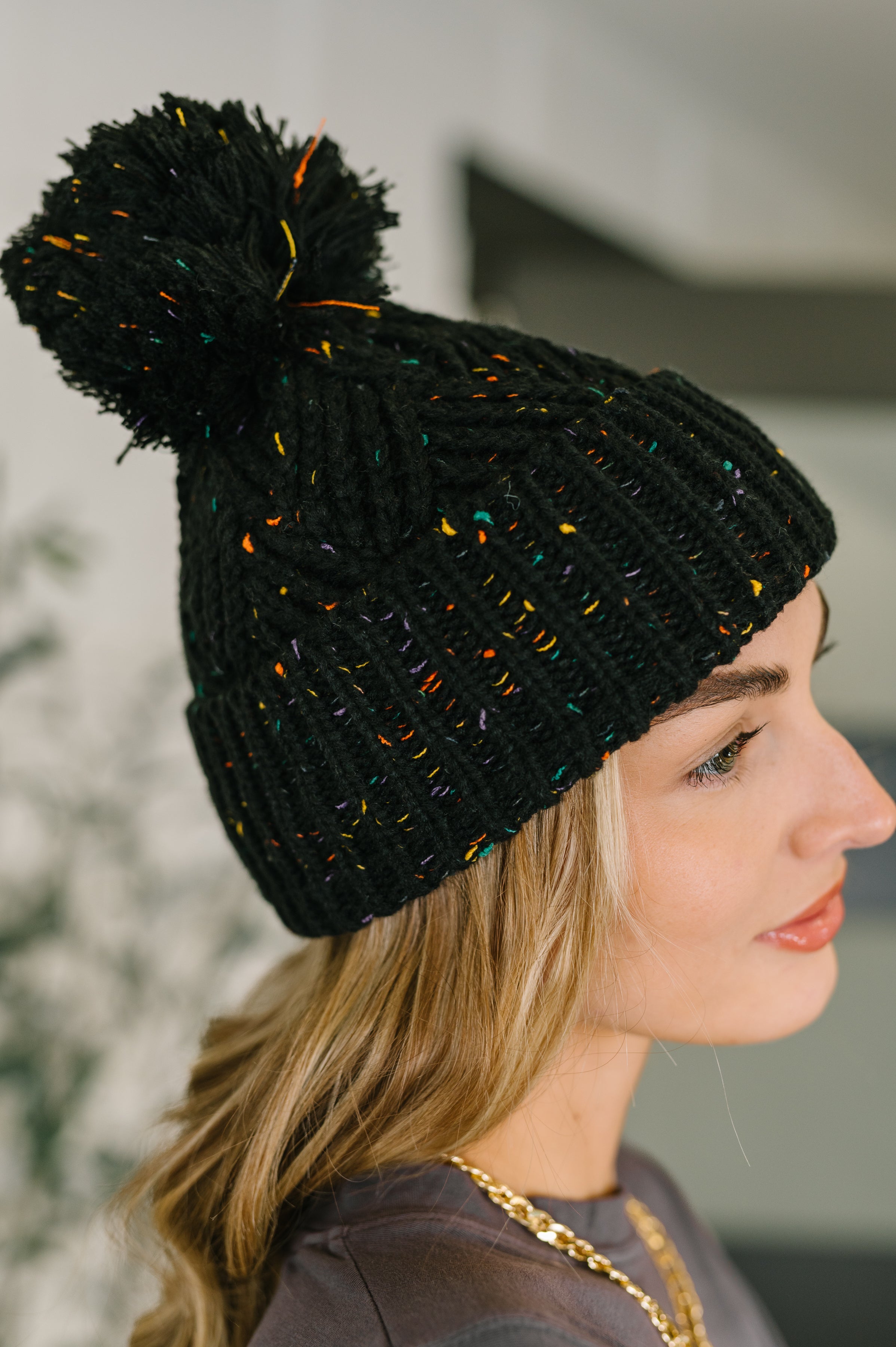 Side profile of woman wearing black knit beanie with colorful flecks and large pom pom on top indoors