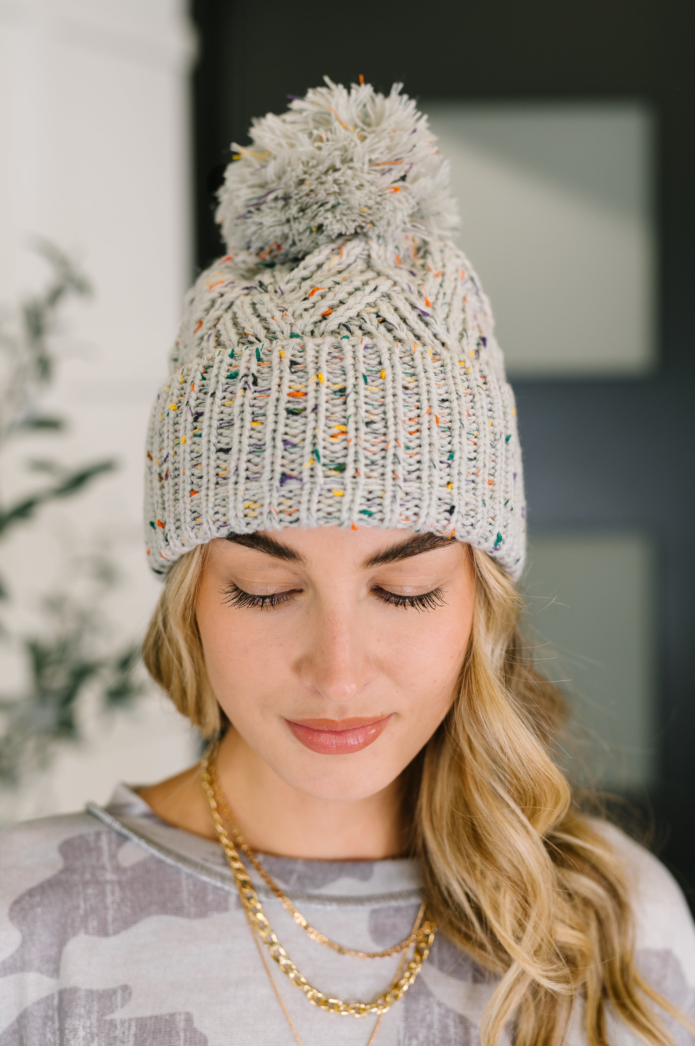 Woman with blonde hair wearing grey knit pom pom beanie with multicolor flecks and layered gold chains indoors
