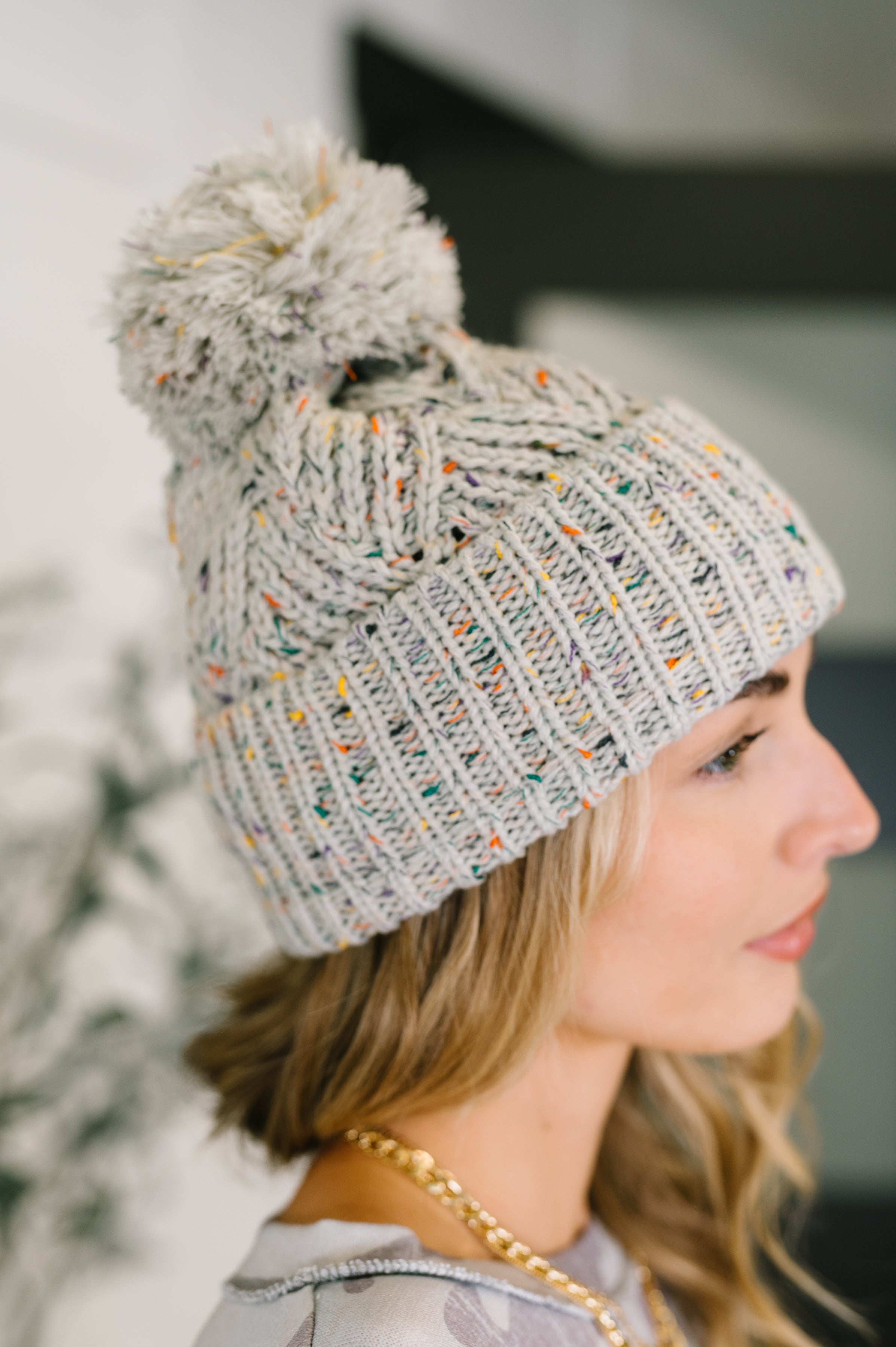 Side view of woman wearing grey knit beanie with colorful specks and large pom pom indoors