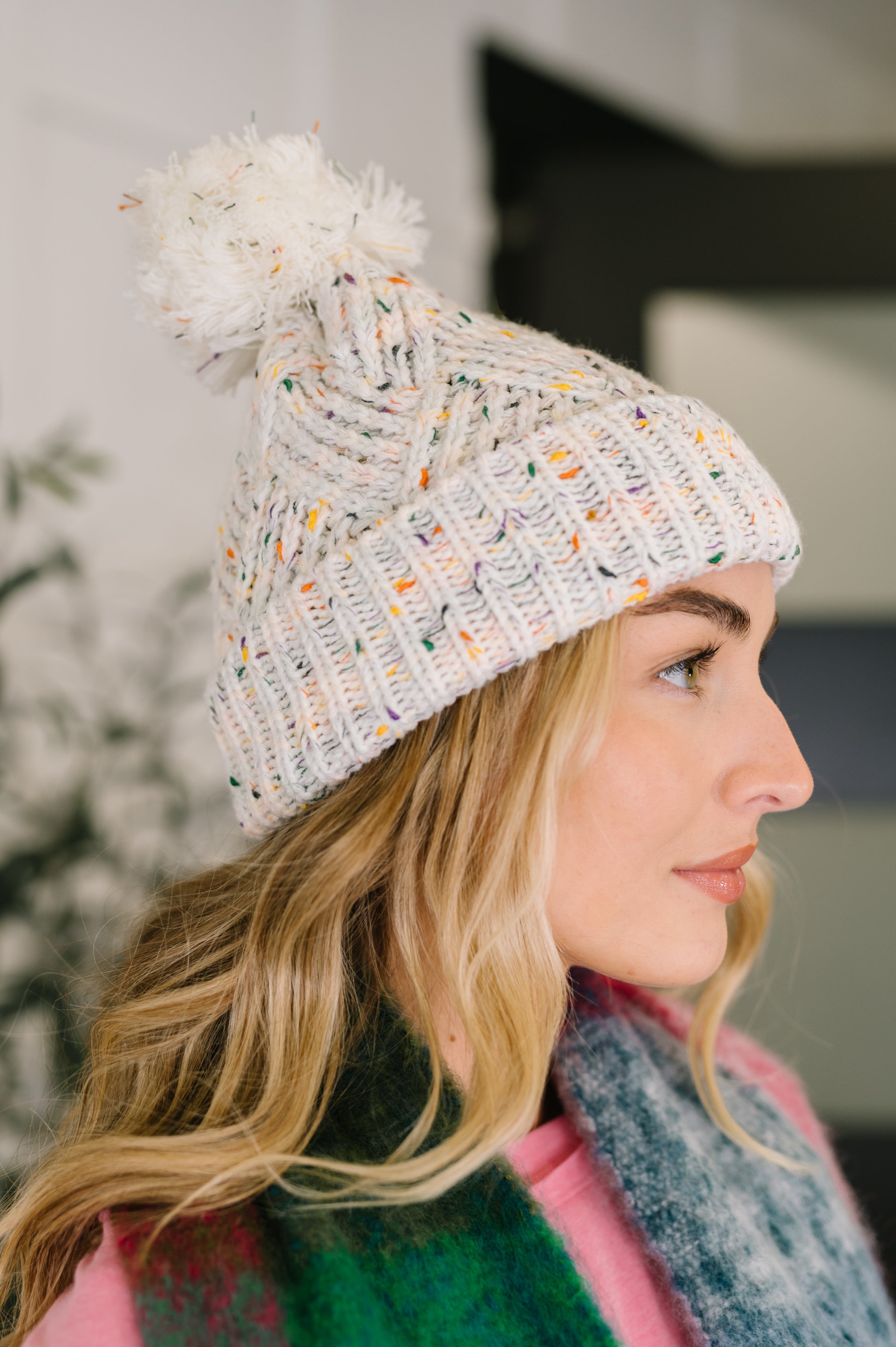 Side profile of woman wearing ivory knit beanie with colorful flecks and fluffy pom pom indoors