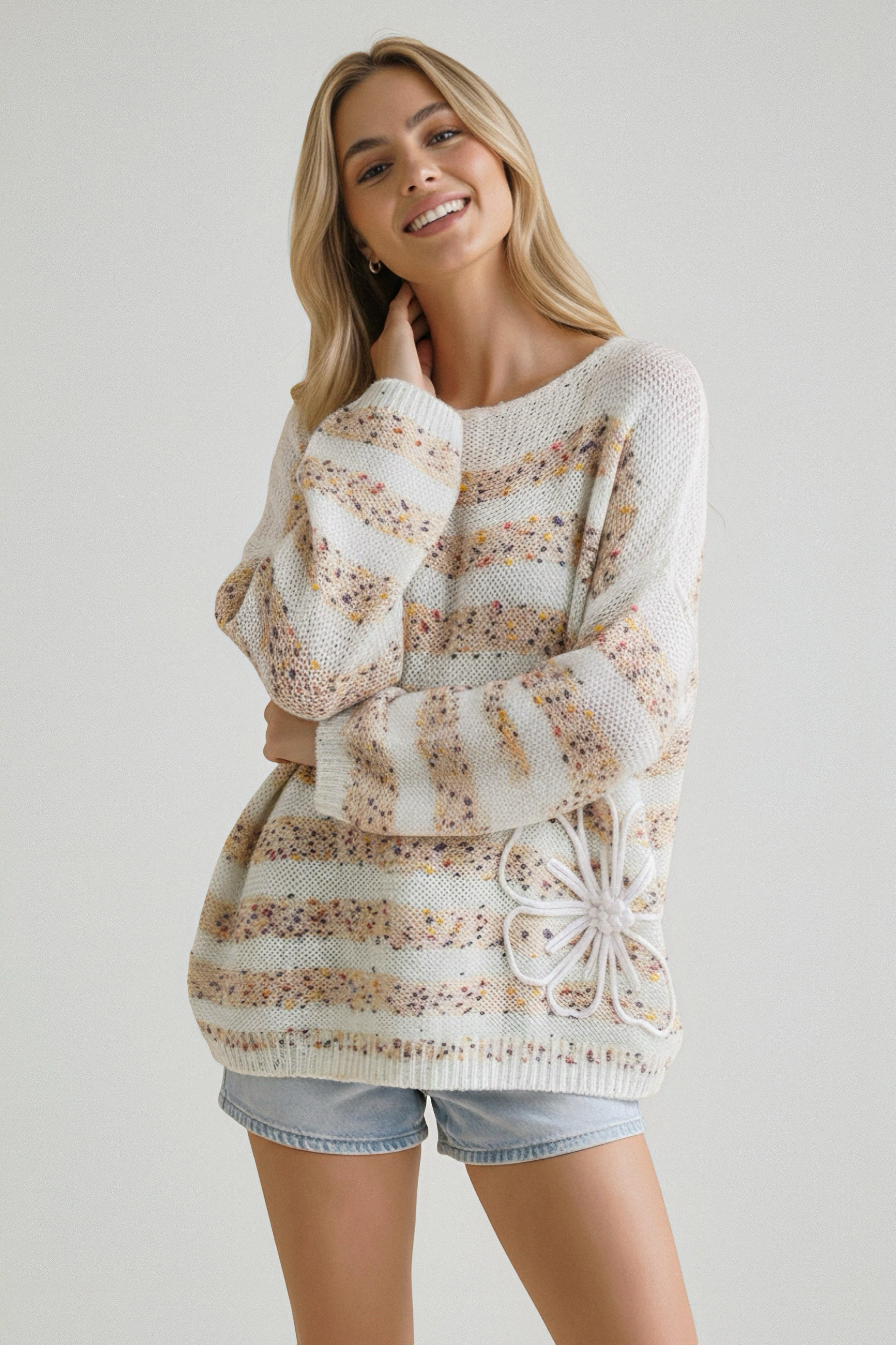 Smiling woman wearing a beige knit sweater with stripe and floral details paired with light denim shorts