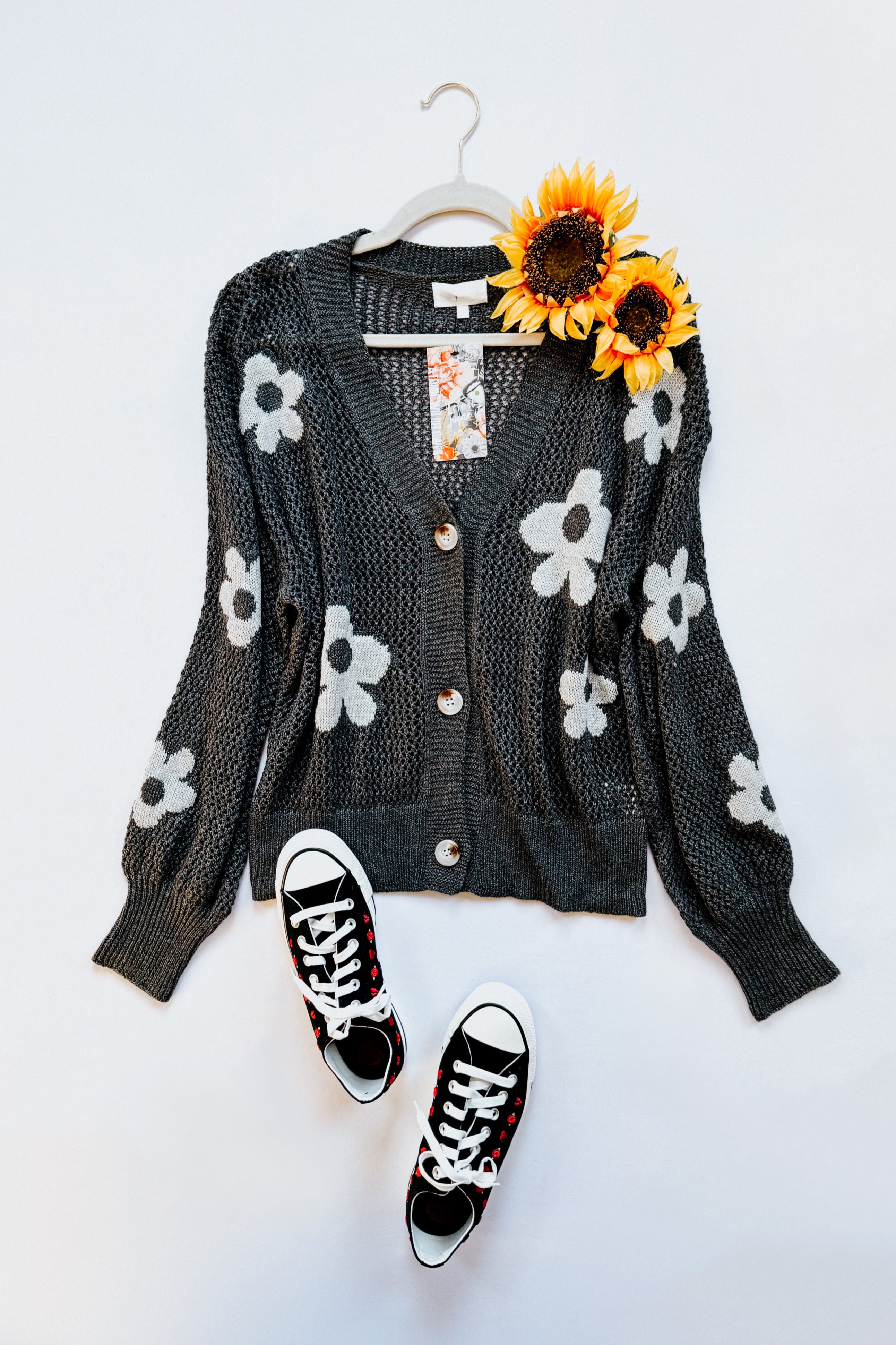 Dark gray floral mesh cardigan with large white flowers and button front laid flat with sunflowers and black sneakers