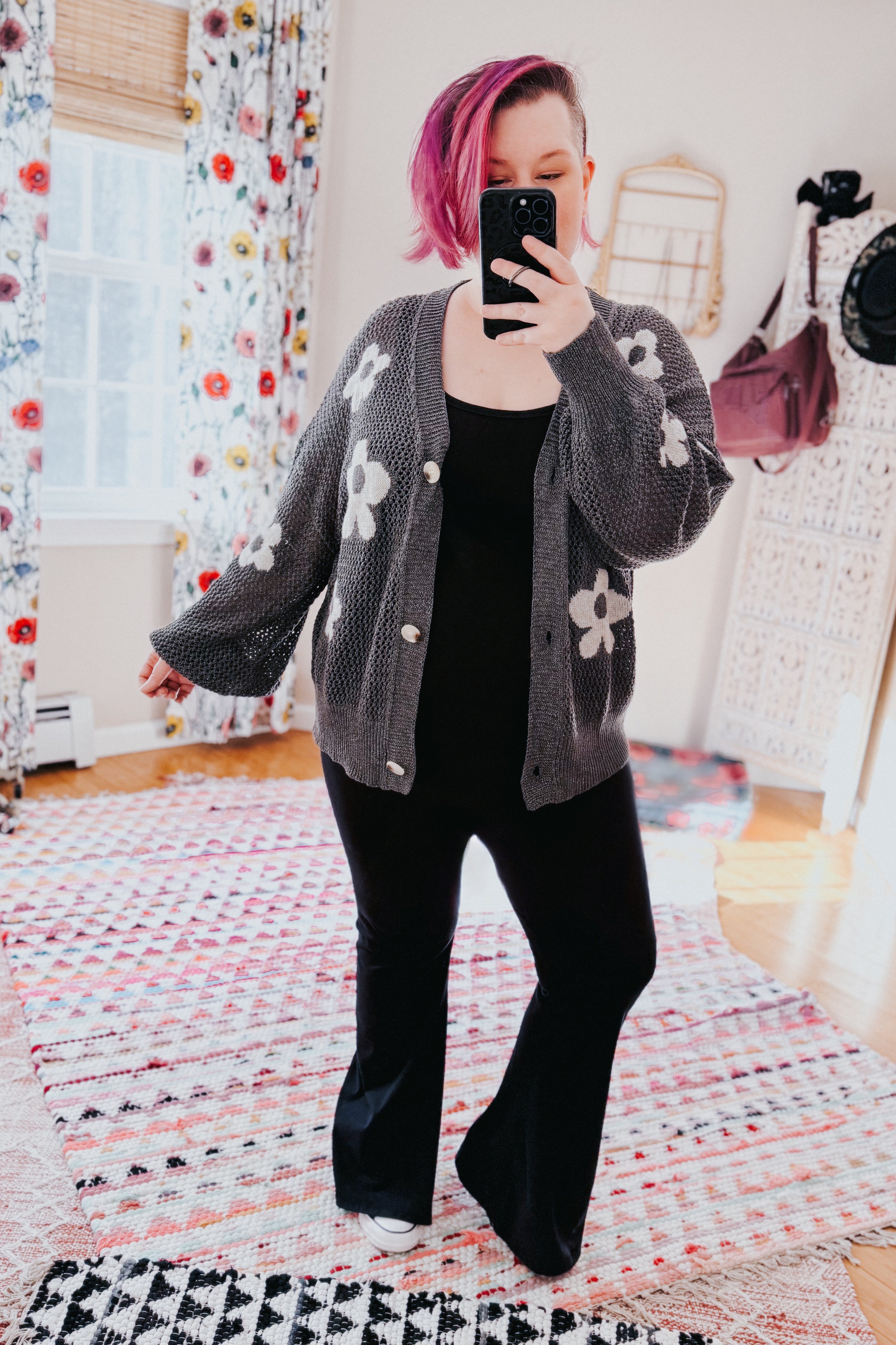 Woman with pink hair taking mirror selfie wearing dark gray floral mesh button cardigan and black outfit in bright room