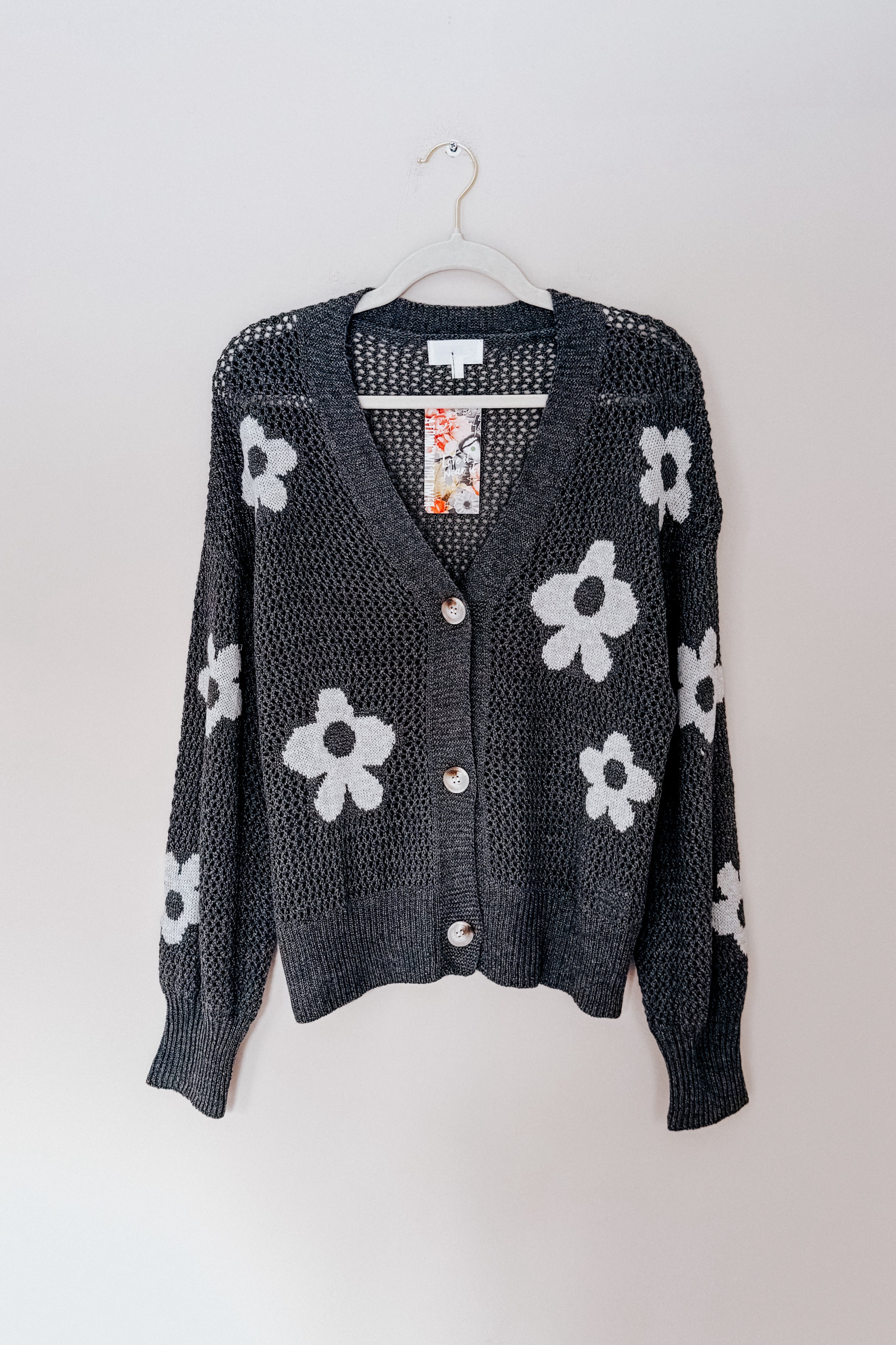 Charcoal gray open-knit cardigan with large white daisy patterns and button closure hanging on white hanger against plain background