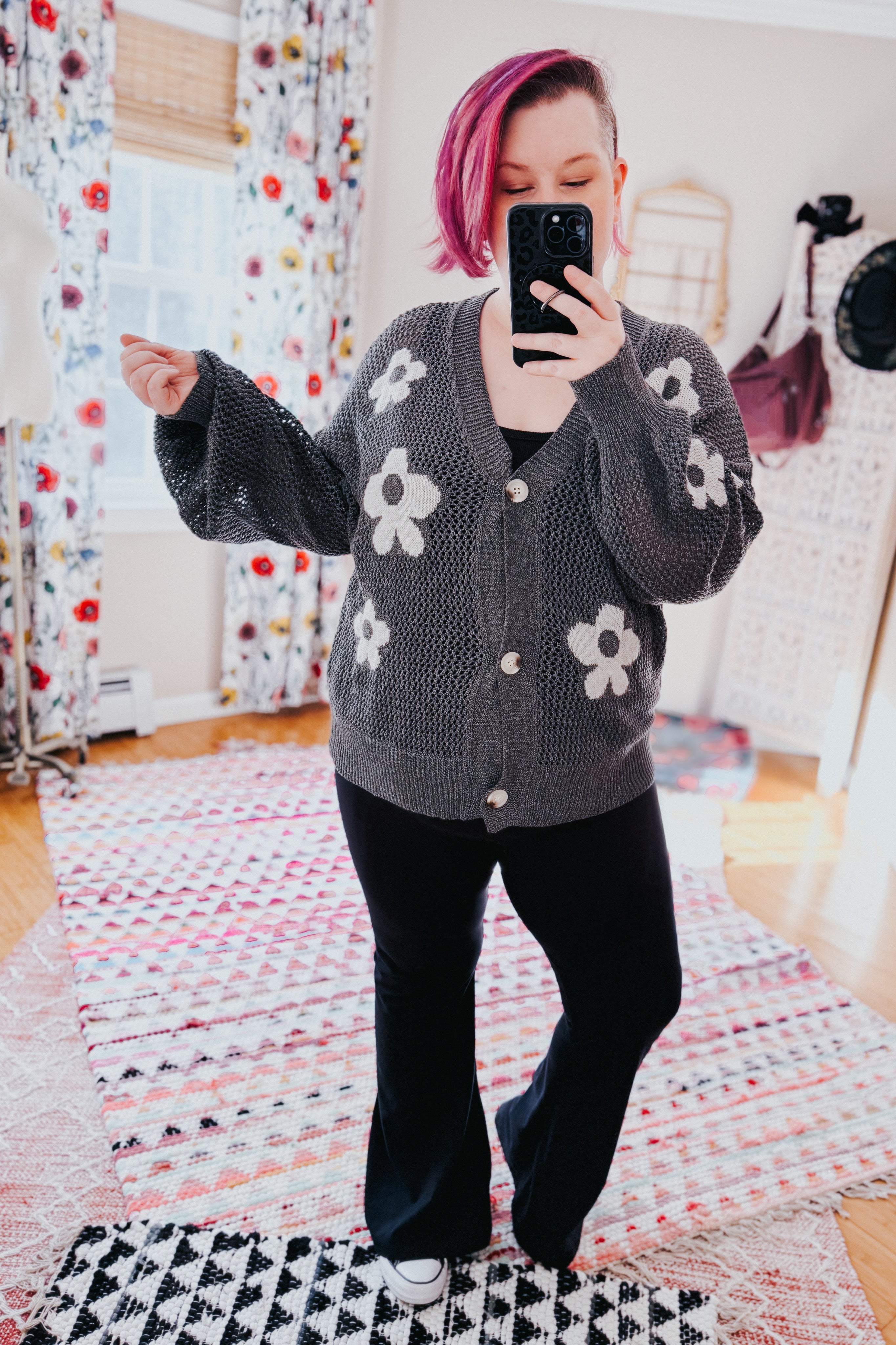 Woman with pink hair taking mirror selfie wearing gray floral mesh cardigan with buttons and black pants in bright room