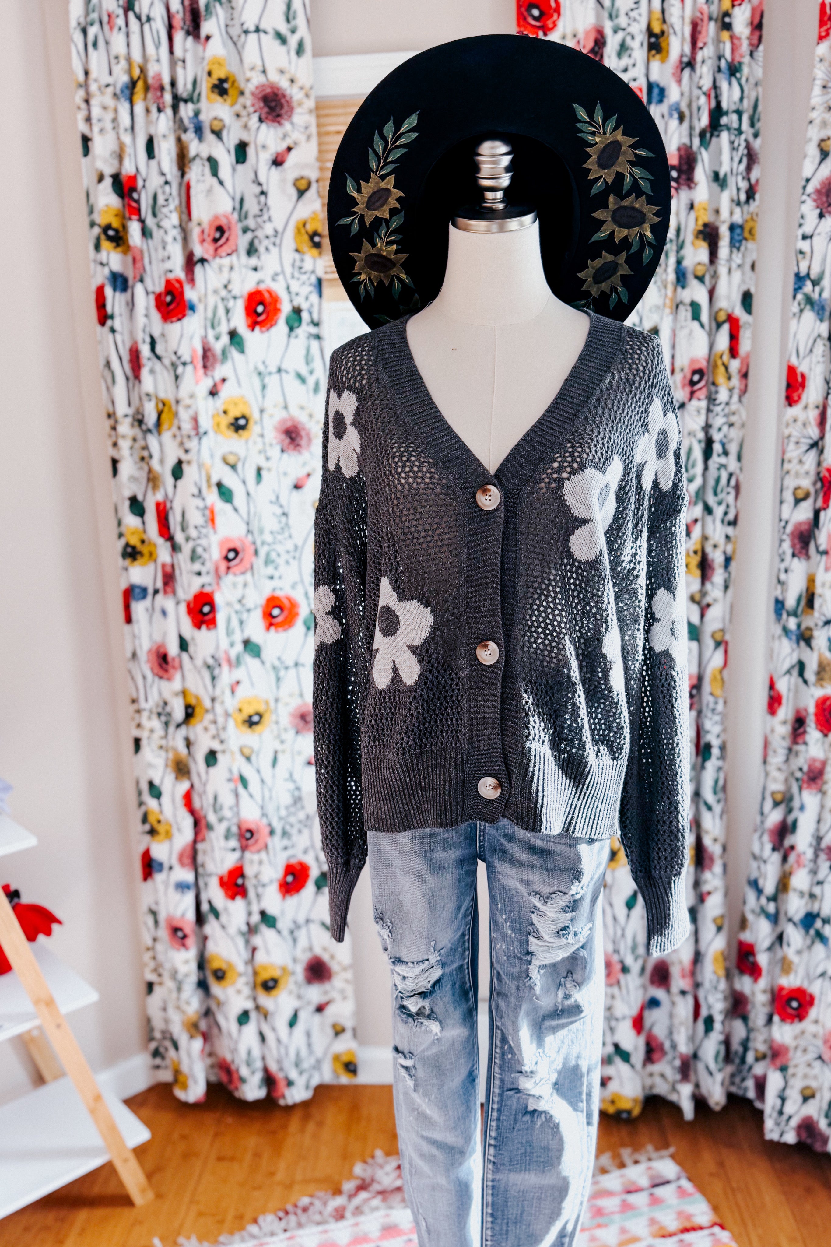 Dark gray mesh cardigan with white daisy patterns and buttons styled on mannequin with black floral hat and distressed jeans
