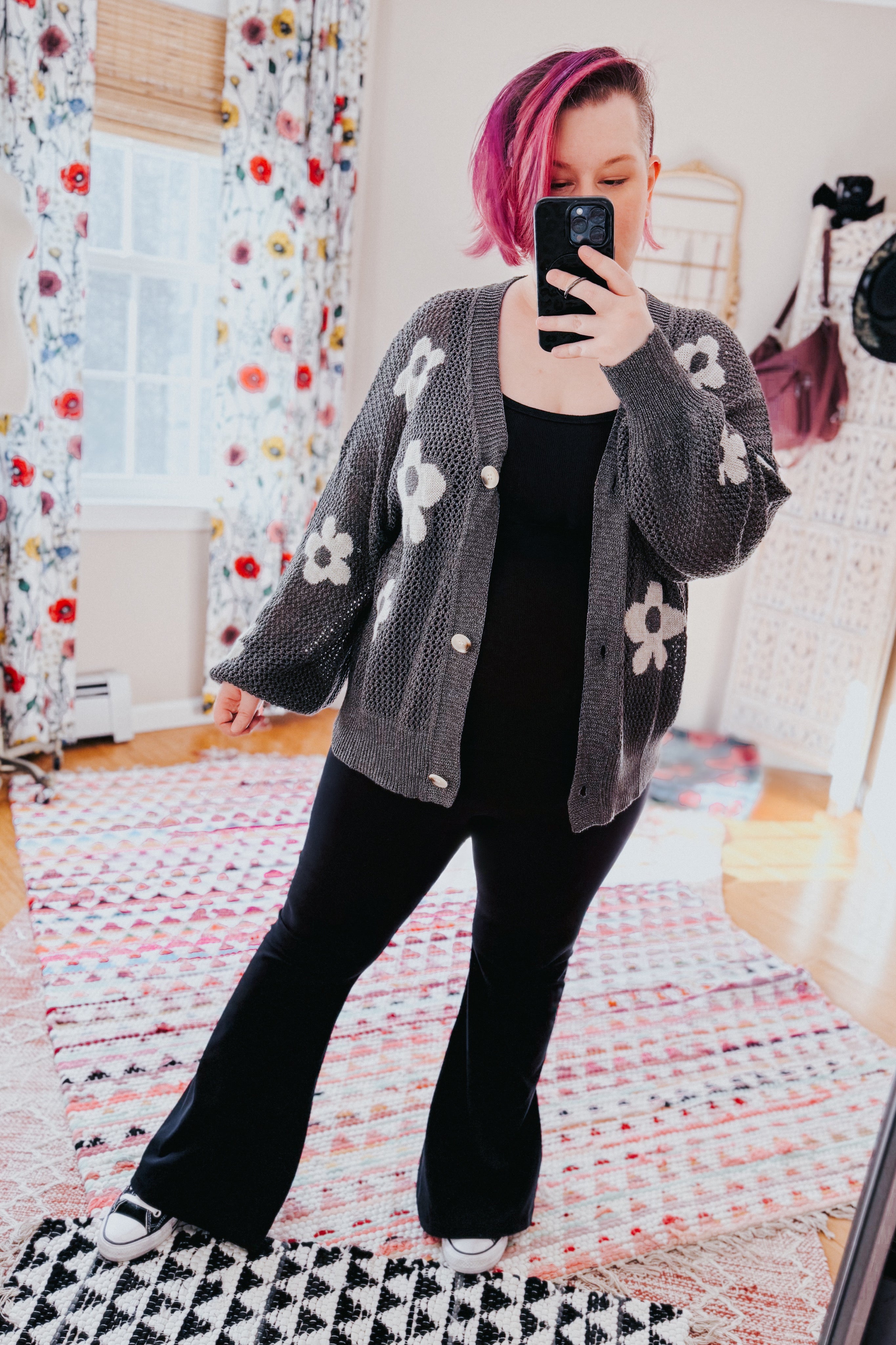 Woman with pink hair taking mirror selfie wearing gray floral button cardigan and black outfit in bright room
