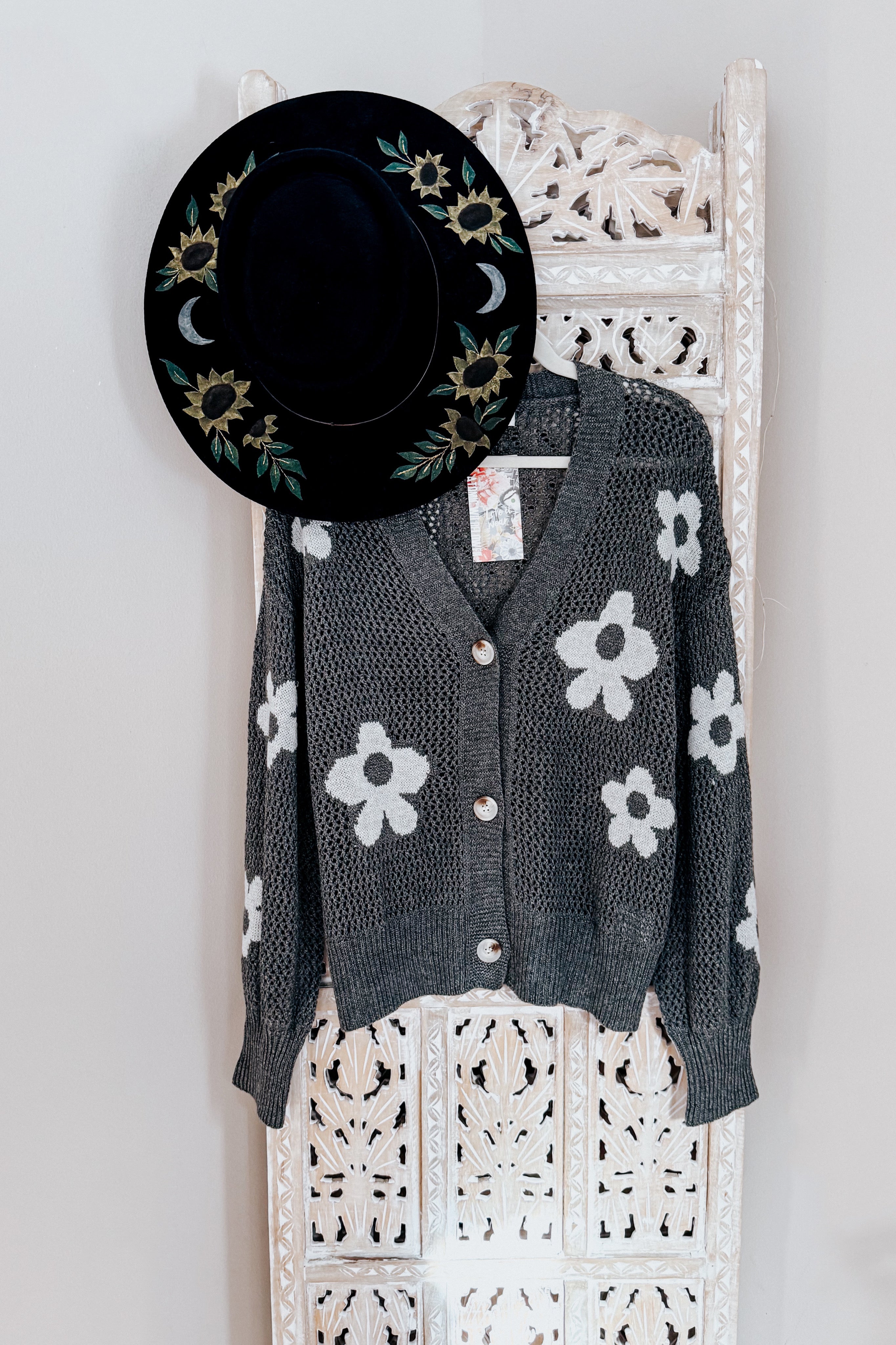 Dark gray knitted cardigan with white daisy motifs and buttons hanging on carved white wooden panel with black floral embroidered hat