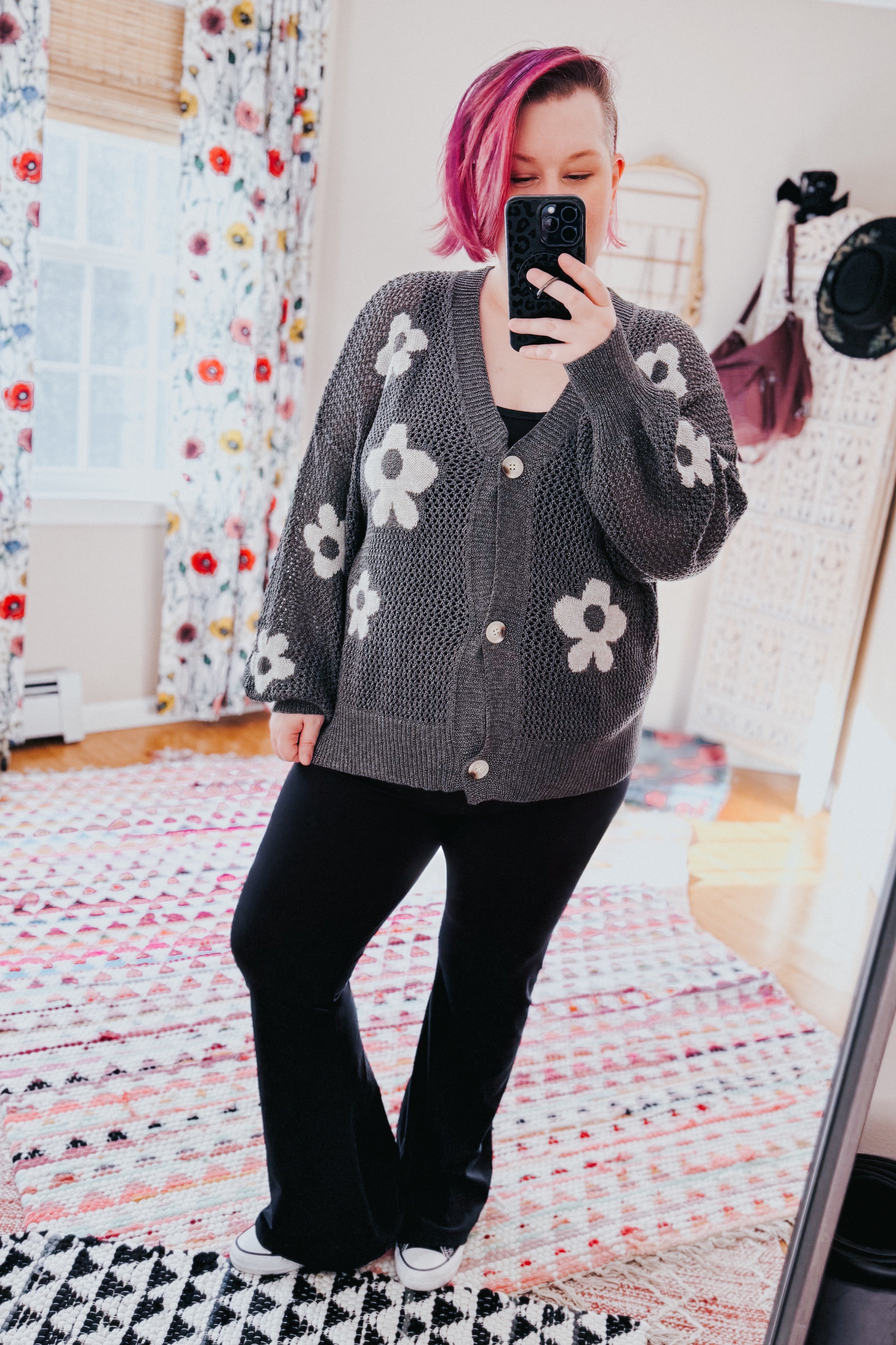 Woman with pink hair taking mirror selfie wearing gray floral knit button cardigan and black flared pants in bright room