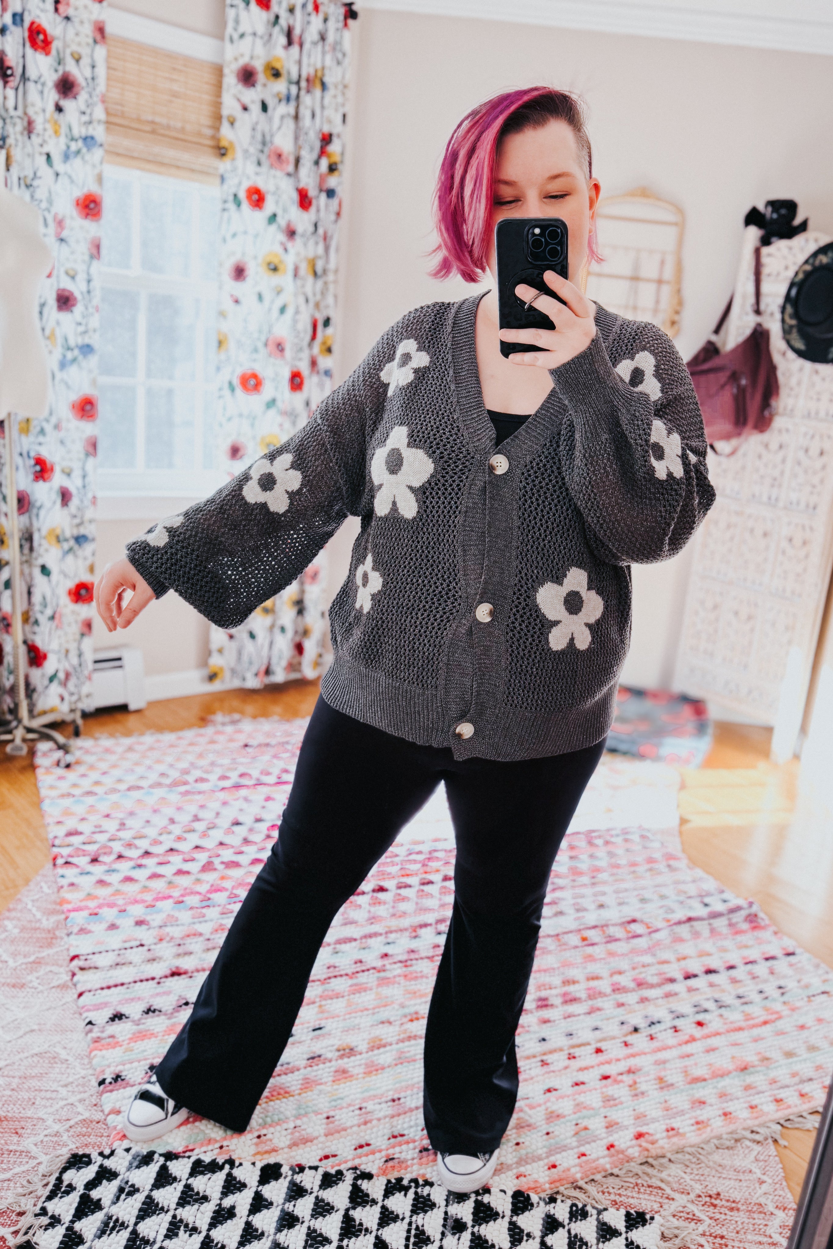 Woman with pink hair taking selfie wearing dark gray mesh cardigan with large white floral patterns and black pants in cozy room