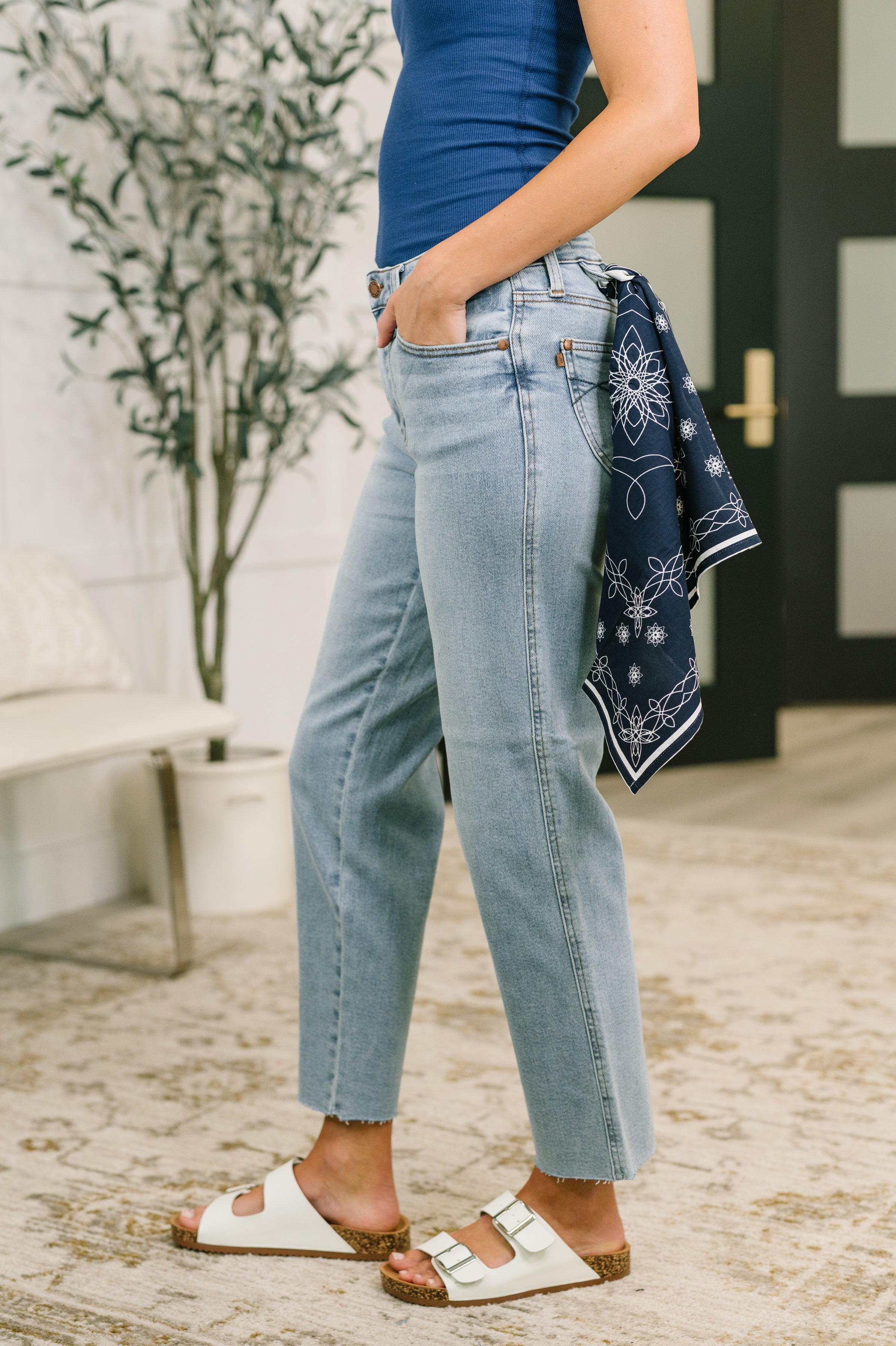 Side view of woman wearing light blue mid rise straight jeans with navy bandana loop and white sandals indoors