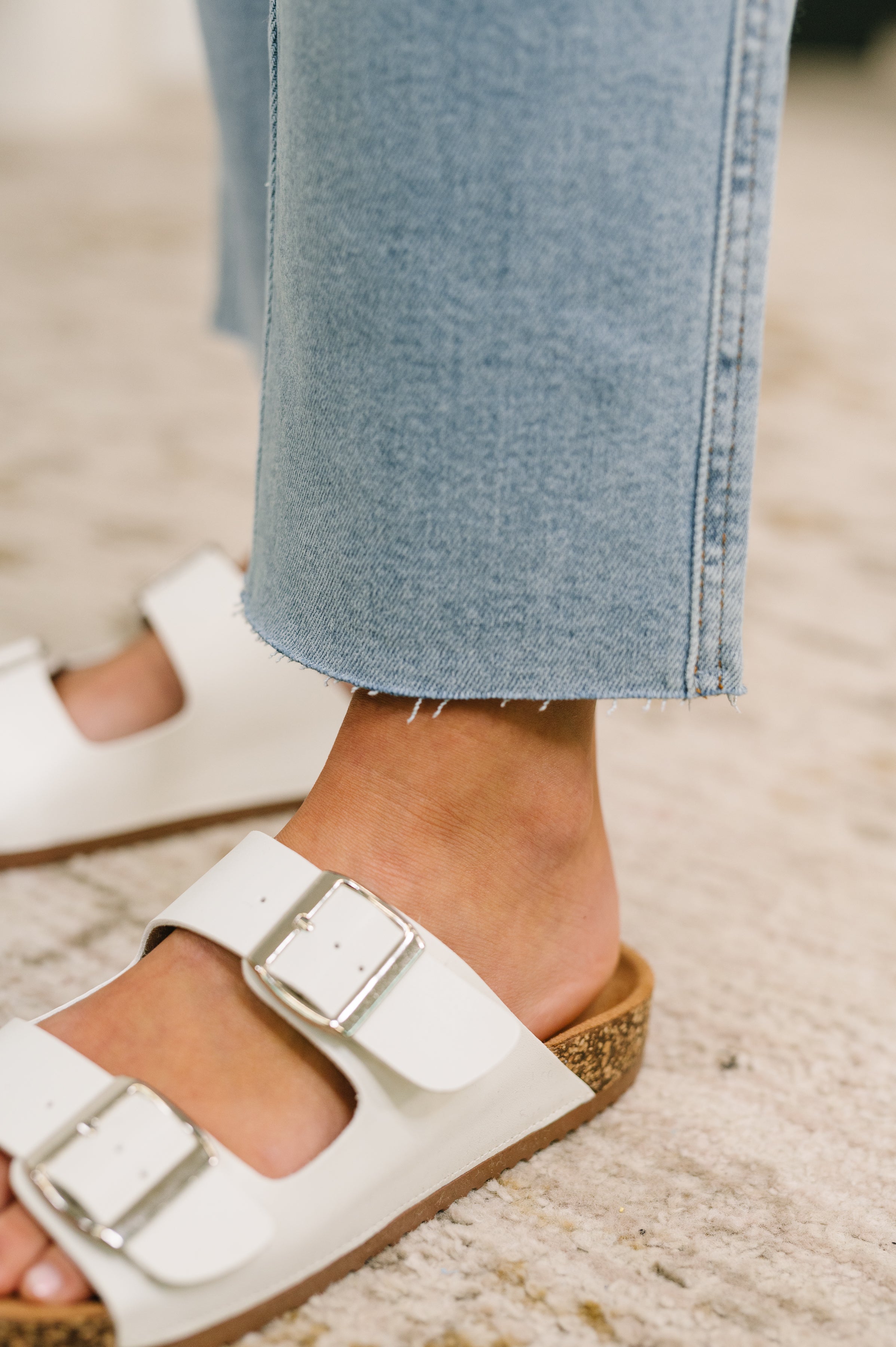 Close-up of ankle-length blue straight leg jeans paired with white buckle sandals on neutral rug background