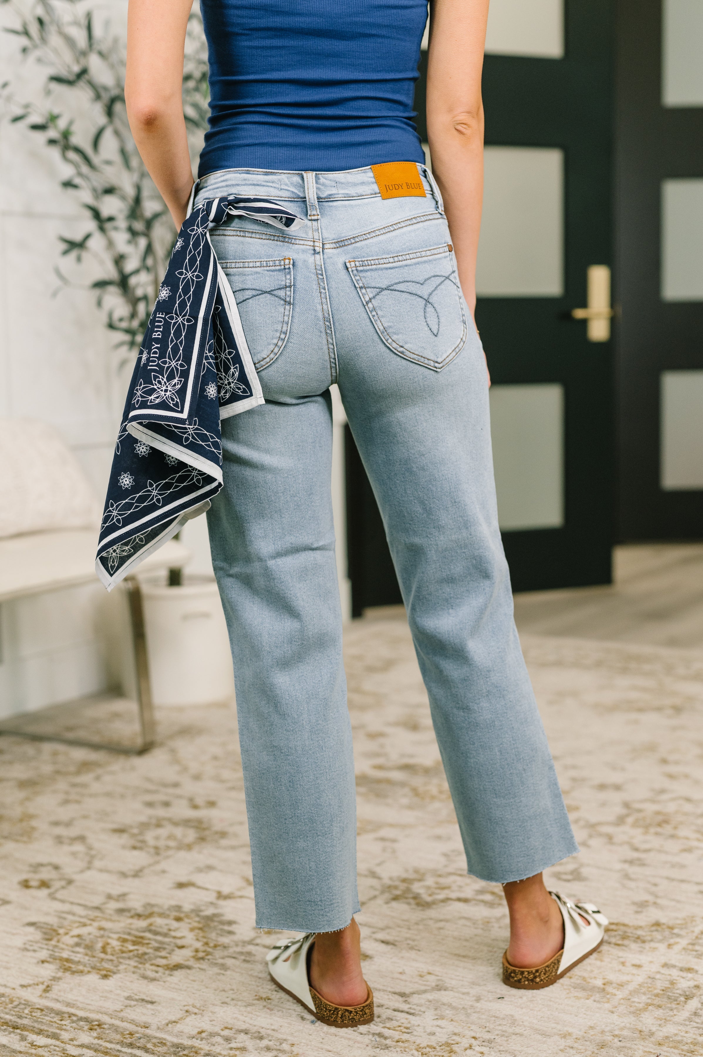 Back view of woman wearing light blue mid rise straight jeans with bandana loop and white slip-on sandals indoors