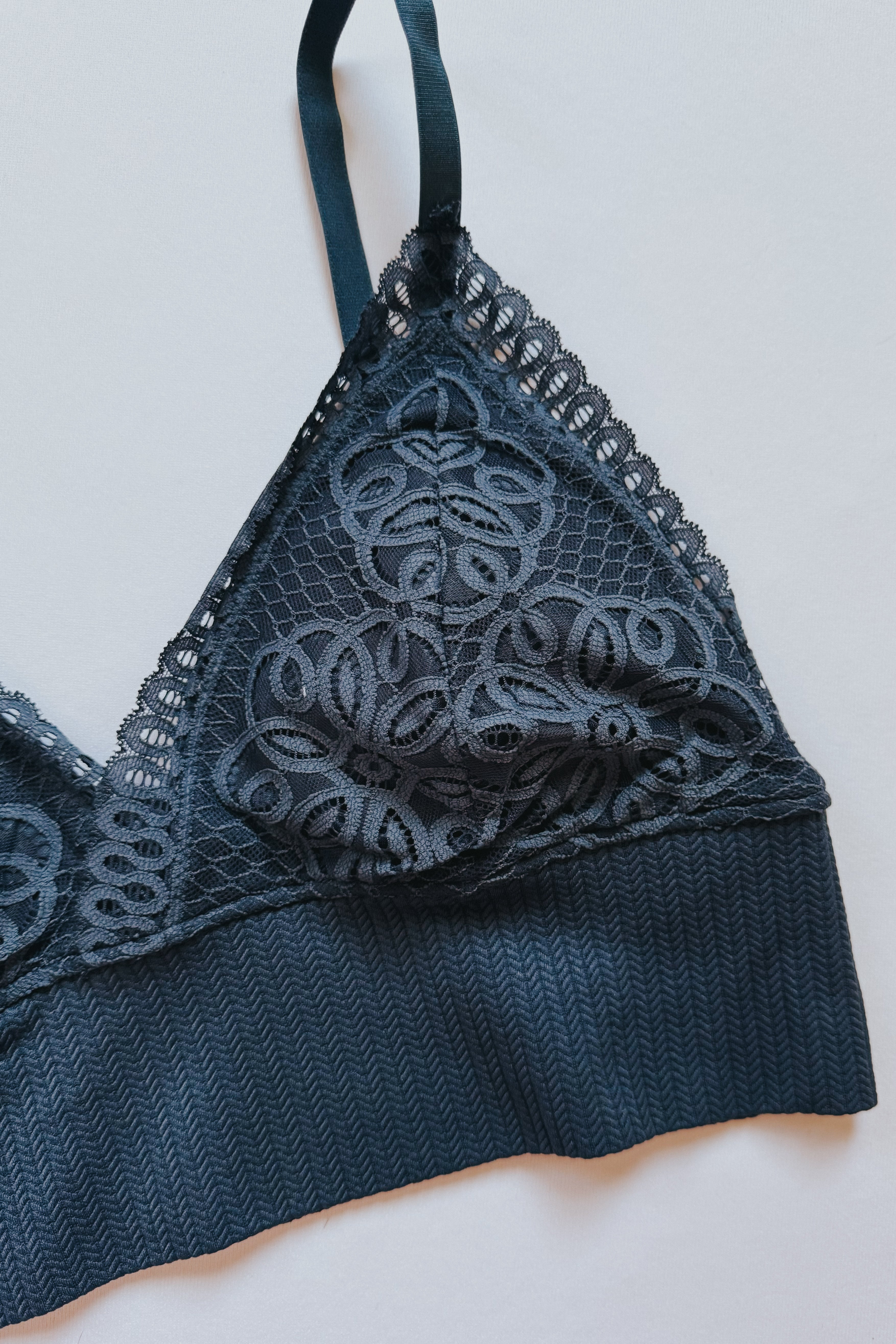 Close-up of dusty blue lace bralette with floral mesh patterns and ribbed band on pale neutral background