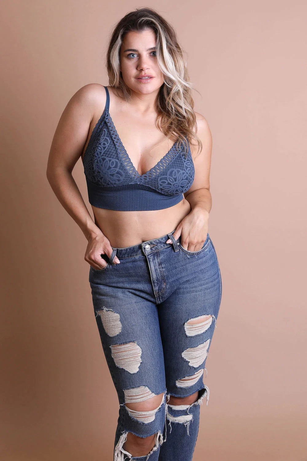 Curvy woman wearing dusty blue lace bralette with geometric design and high-rise distressed denim jeans against beige backdrop