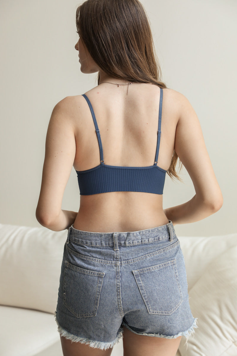 Woman seen from behind wearing dusty blue ribbed bralette with adjustable straps and high-waisted denim shorts indoors